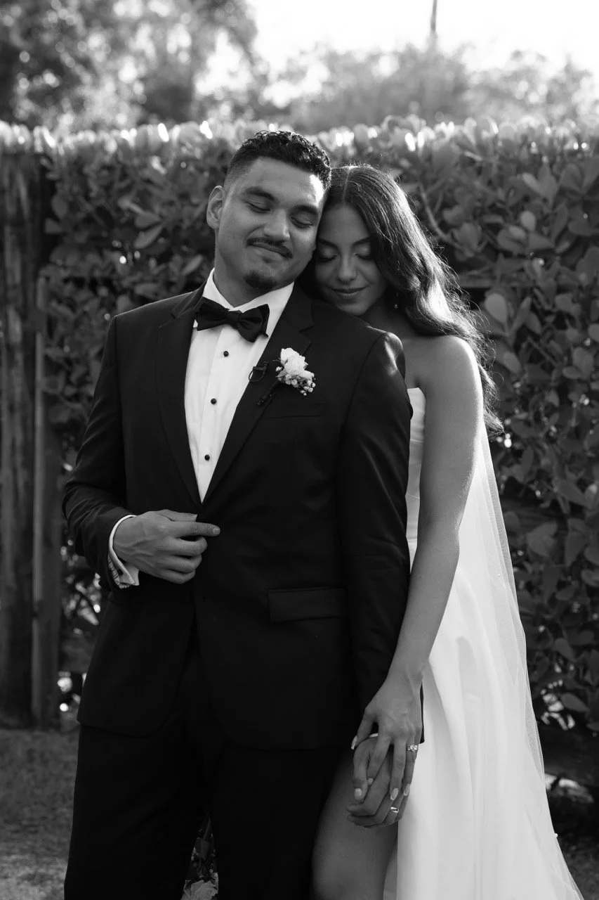 A black and white photo of a couple dressed in wedding attire, standing outdoors with greenery in the background. The man is wearing a tuxedo with a bow tie and a boutonniere, and the woman is in a strapless wedding dress with her hand on his arm.