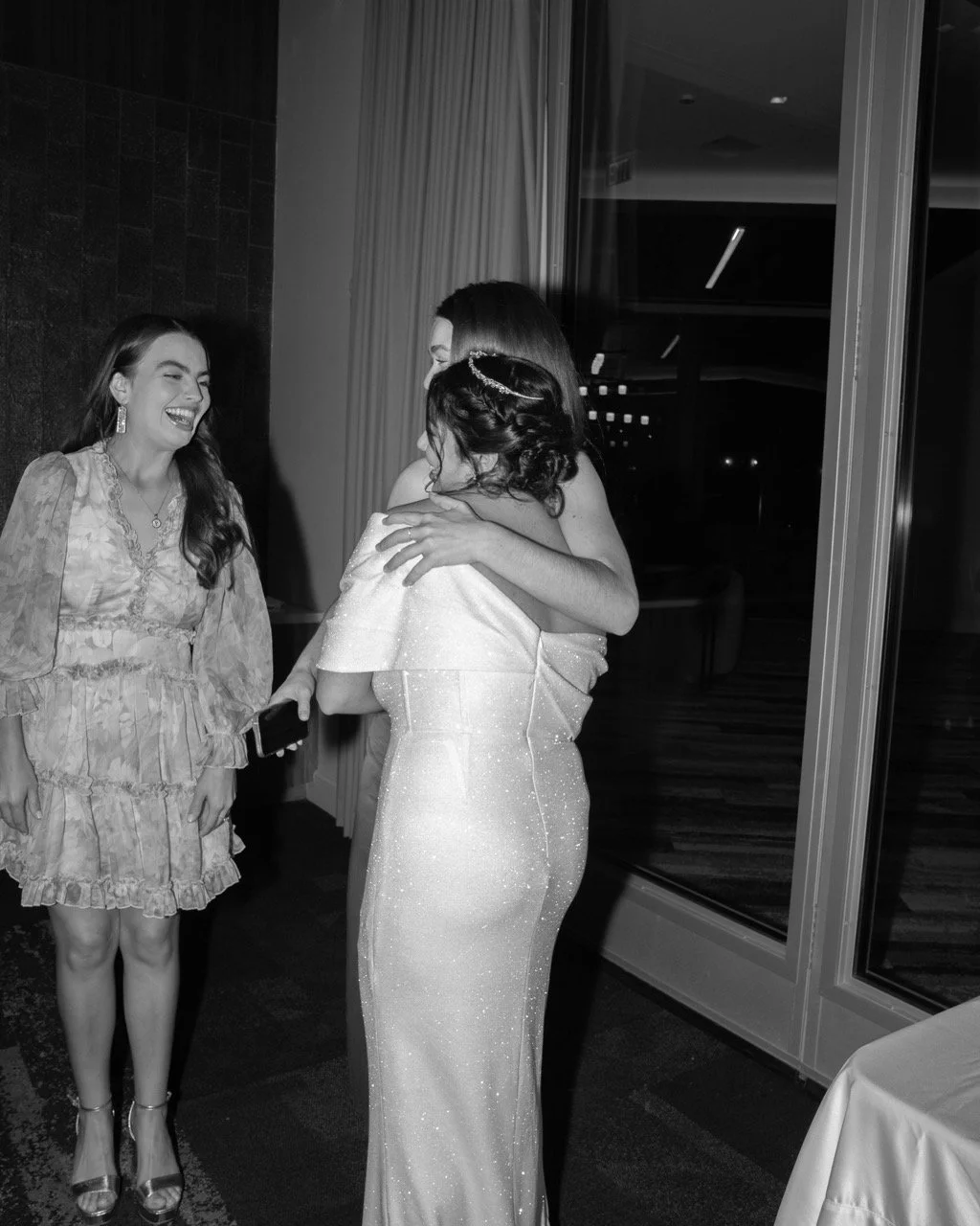 Three women at an indoor event, one in a shiny gown hugging another, the third smiling and holding a phone.