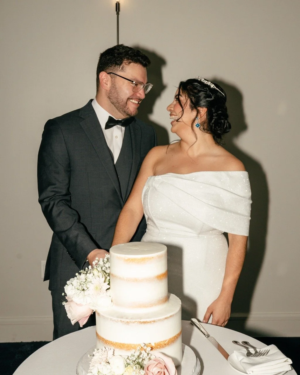 Bride and groom smiling at each other during wedding cake cutting, with a multi-layered cake decorated with flowers in front of them.