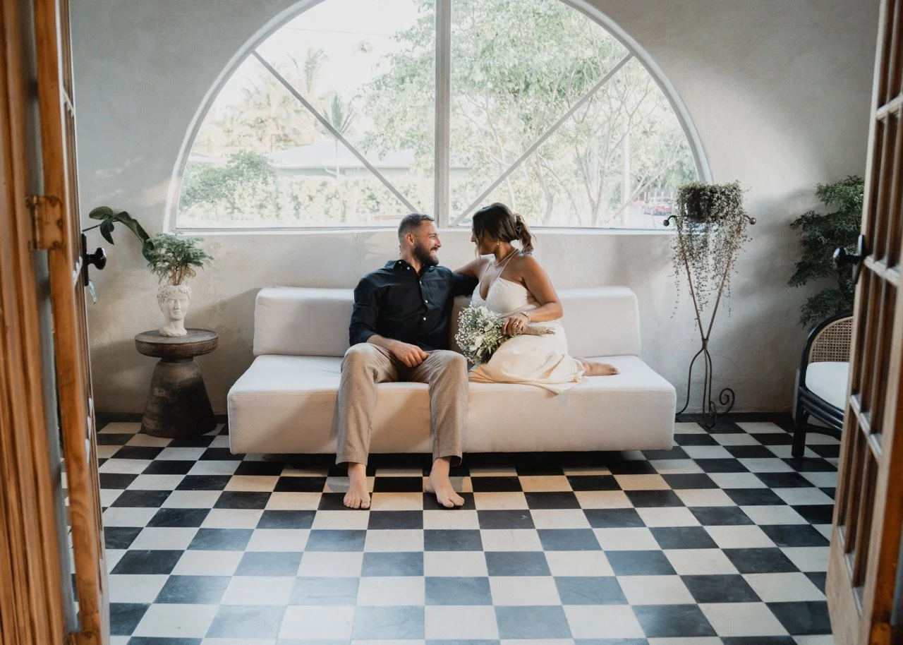 A smiling couple sitting on a white couch inside a room with a checkered black and white floor, large arched window, and potted plants, sharing an intimate moment.