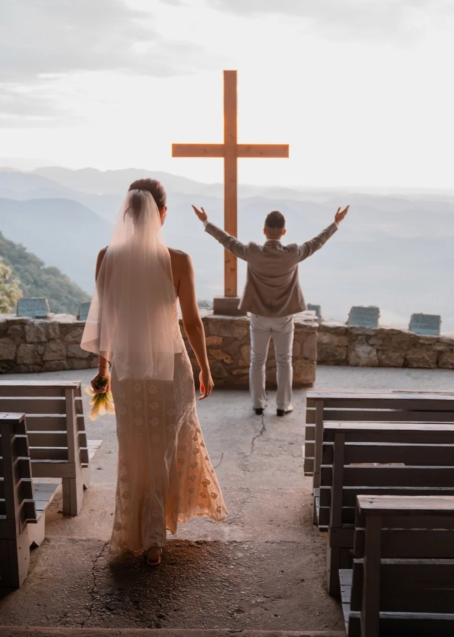 A bride walking down the aisle towards a man with arms raised in front of a large wooden cross overlooking mountains and a cloudy sky.