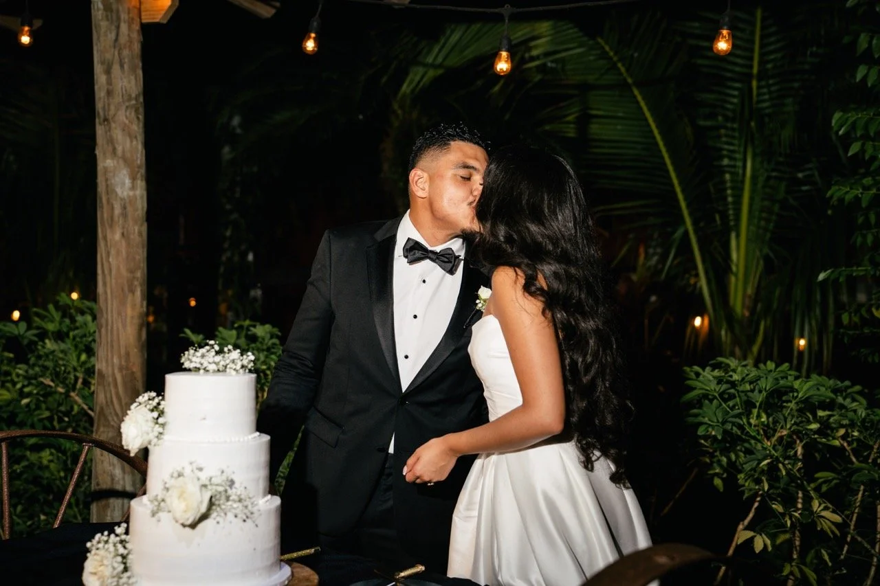 A newlywed couple shares a kiss at their wedding reception; the groom is wearing a black tuxedo with a bow tie, and the bride is in a white wedding dress with long black hair. A three-tiered white wedding cake decorated with white flowers is on the t