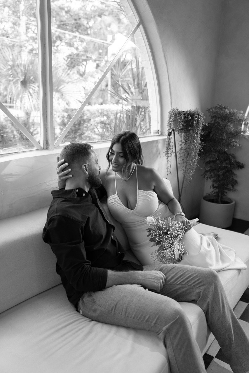 A couple sitting on a sofa in a room with large windows, smiling at each other. The woman is holding a bouquet of flowers and touching the man's face.