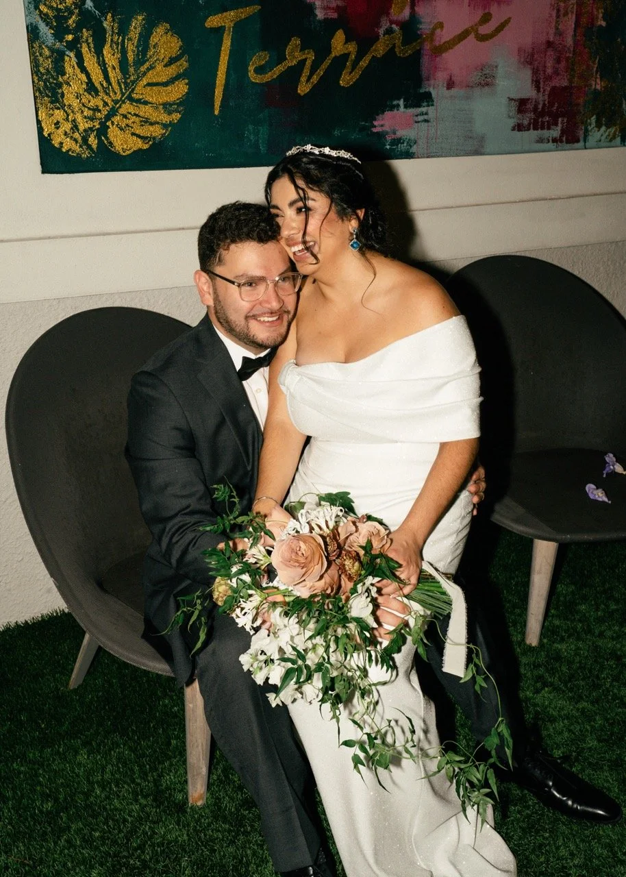 A bride and groom sit together, smiling at each other during their wedding. The bride is dressed in a white off-the-shoulder gown with earrings and a tiara, holding a cascading bouquet of blush roses and white flowers. The groom wears a black tuxedo 