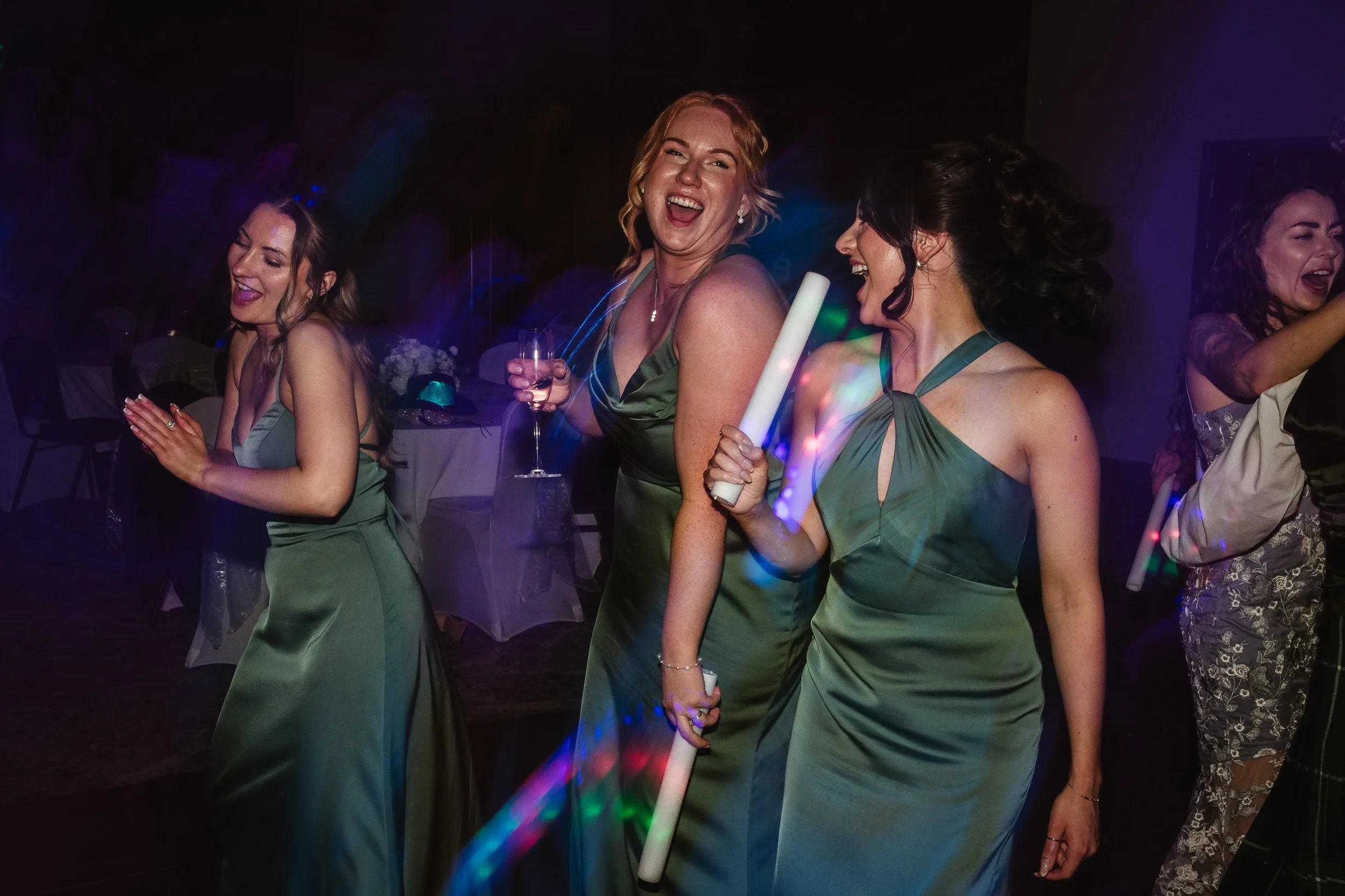 Women in formal dresses dancing and celebrating at a party or wedding reception, with one holding a glow stick.