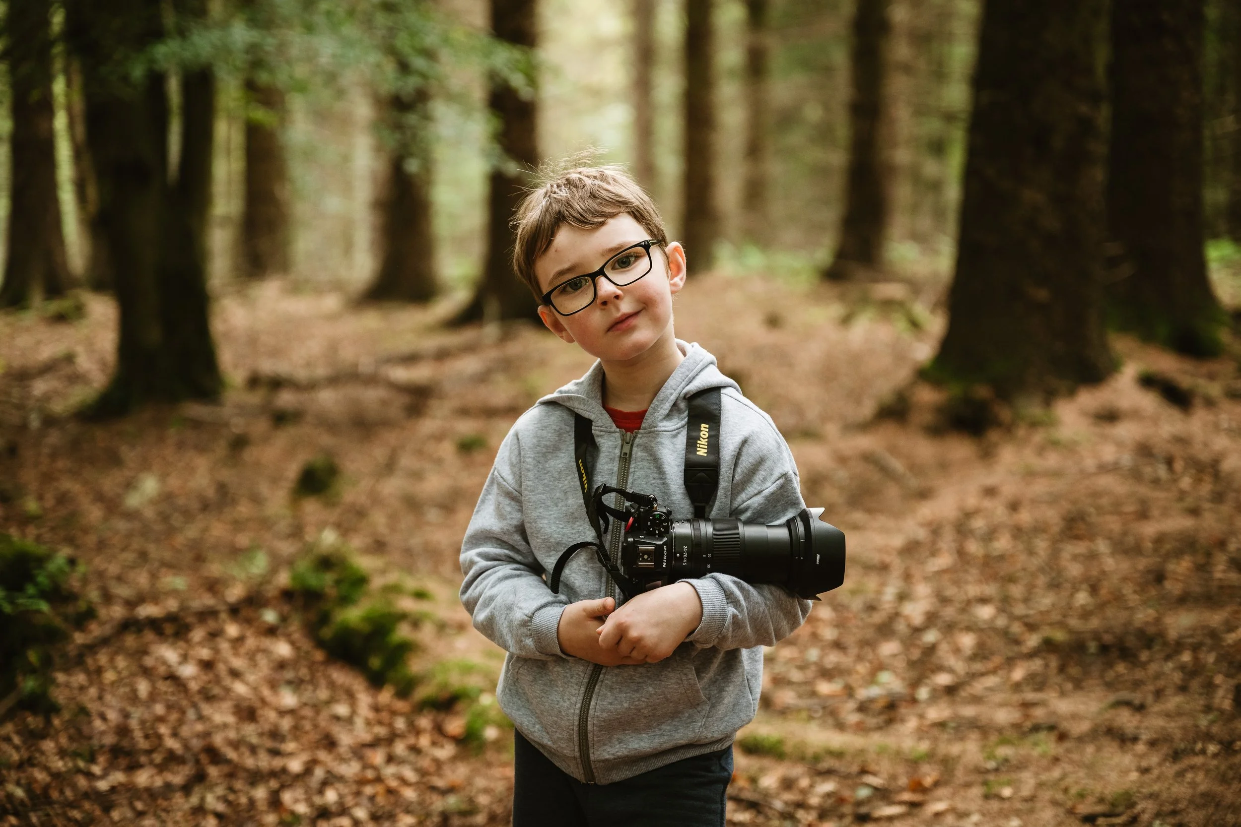Young boy with glasses holding a camera in a forest.