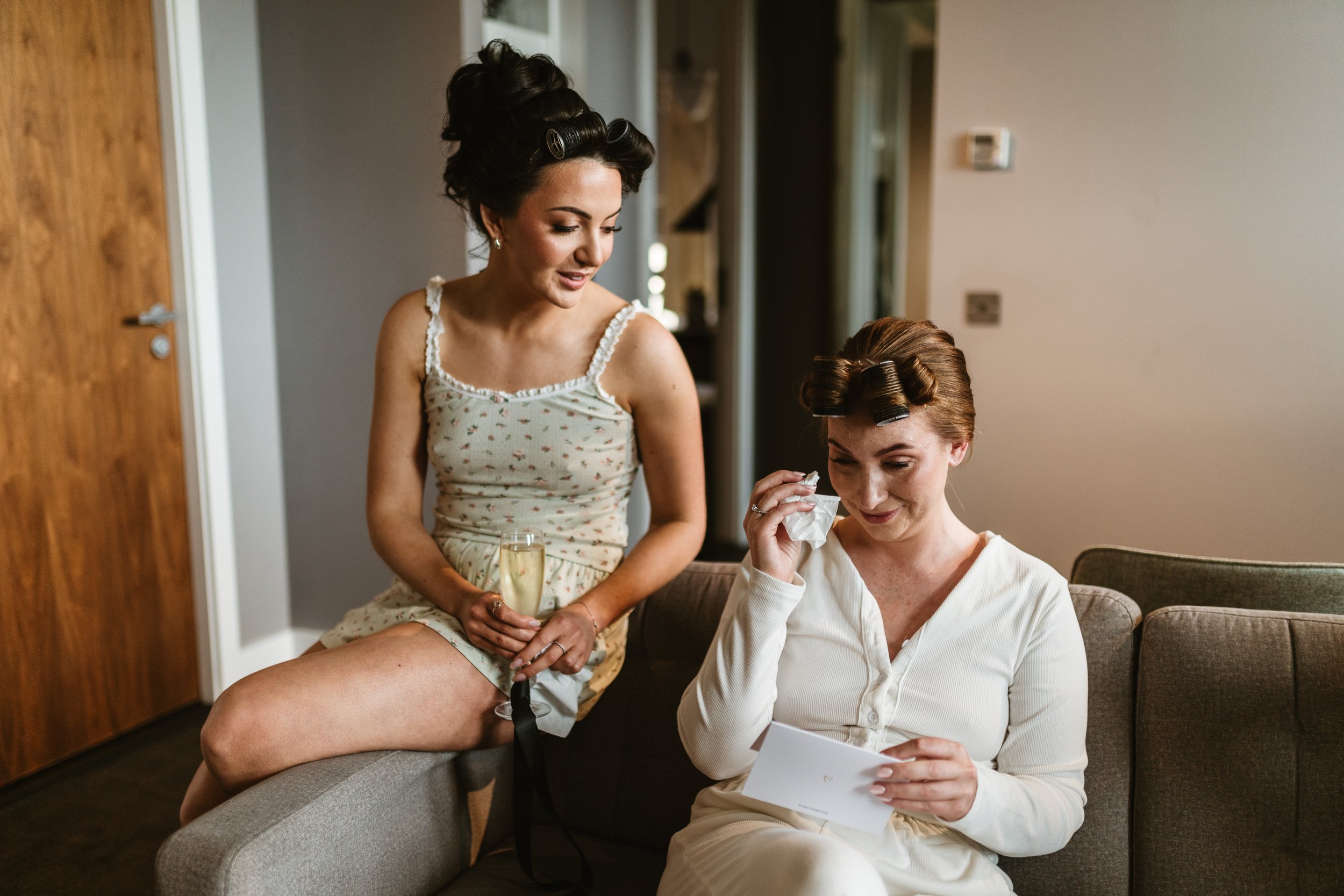 Two women, one sitting on a sofa and the other standing beside her, both with hair rollers, sharing a moment as the woman on the sofa reads a card and the other holds a glass of champagne.