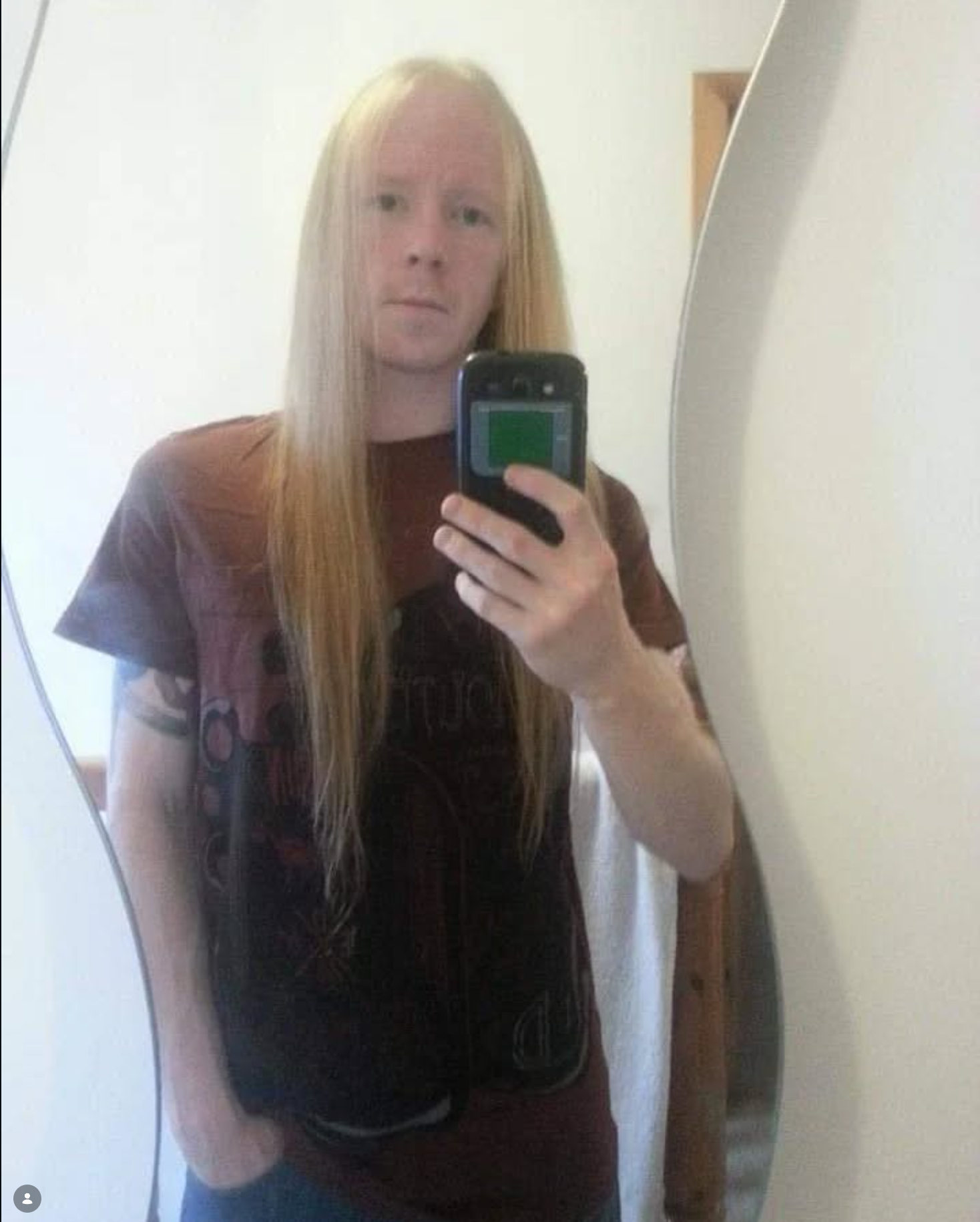 A person with long straight red hair taking a mirror selfie using a mobile phone, wearing a dark T-shirt, with a plain wall in the background.