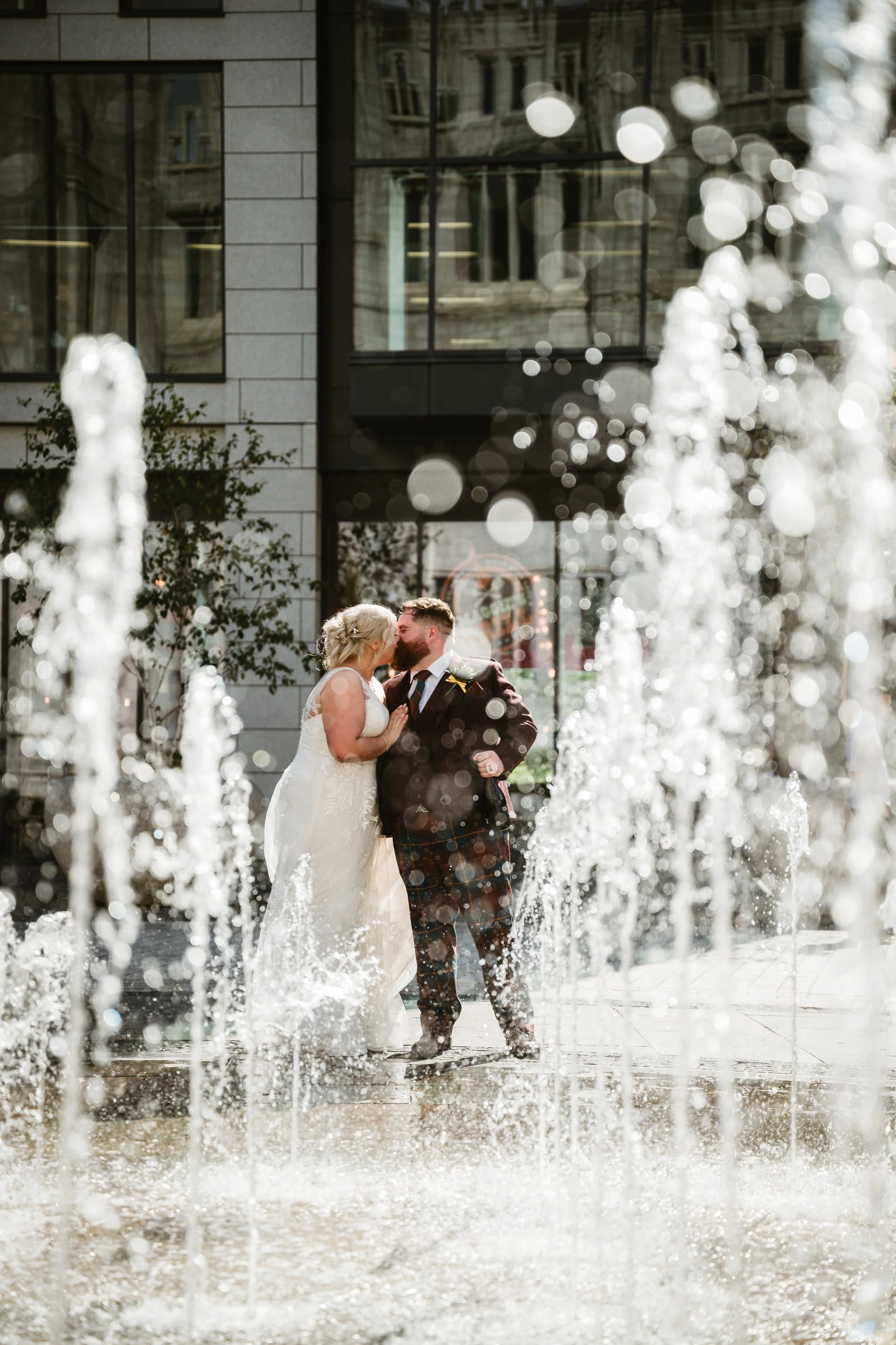 A bride and groom stand kissing in the middle of a water fountain, with tall buildings and windows in the background, during a wedding photo shoot.