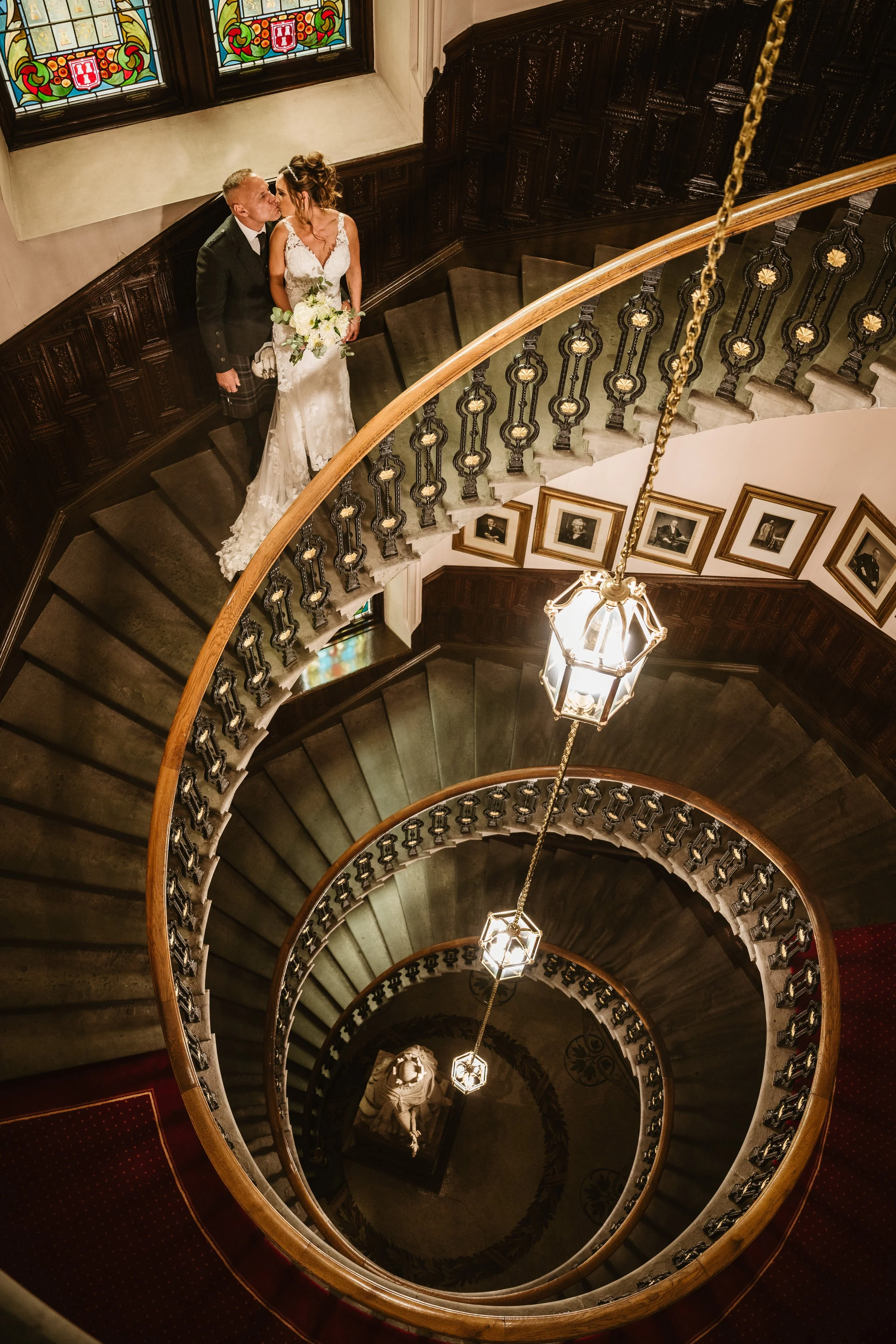 A bride and groom stand on a spiral staircase, sharing a kiss inside a historic building with stained glass windows and framed black-and-white photographs on the walls, illuminated by hanging lanterns.