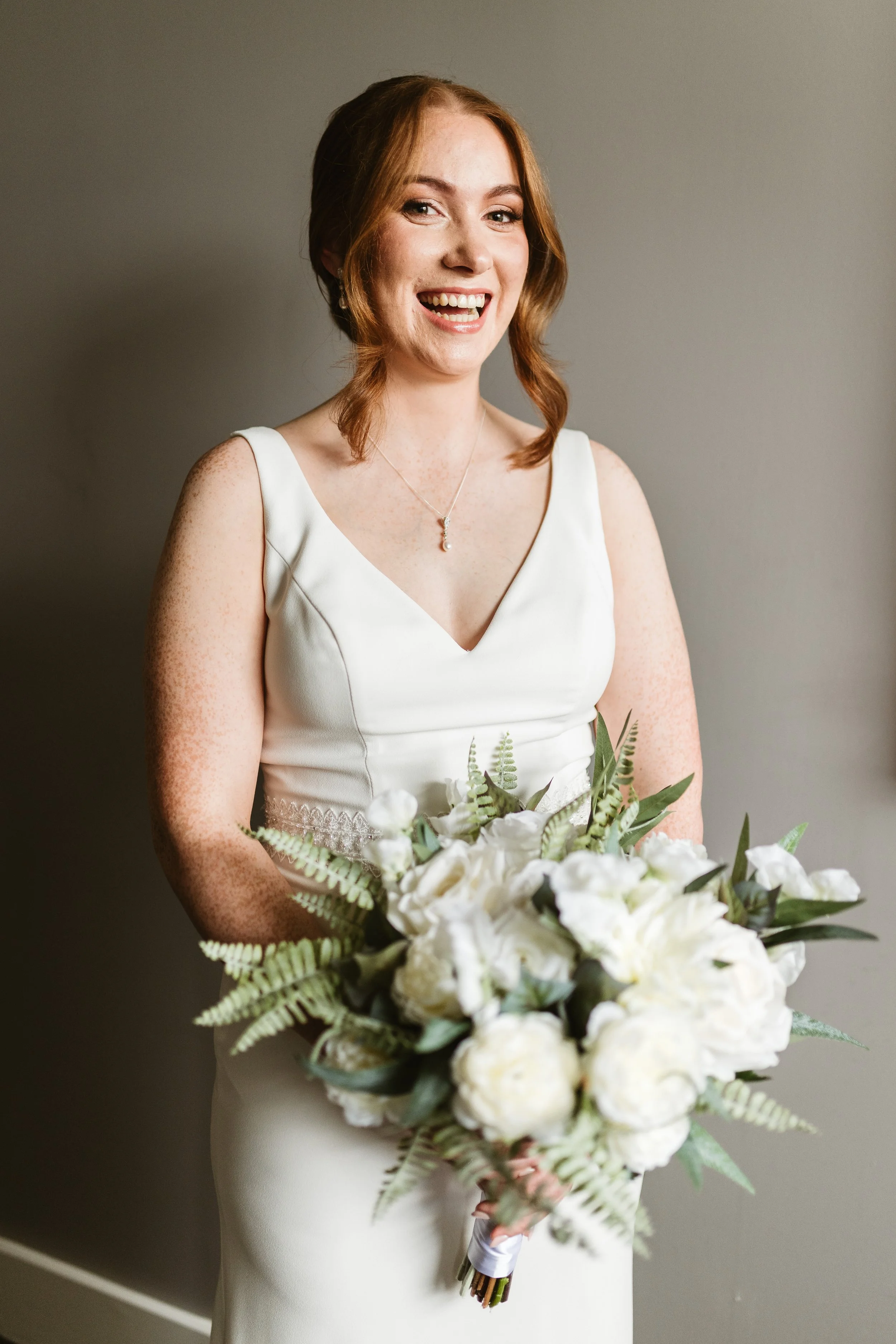 A smiling bride in a white wedding dress holding a large white floral bouquet with greenery, standing against a plain background.