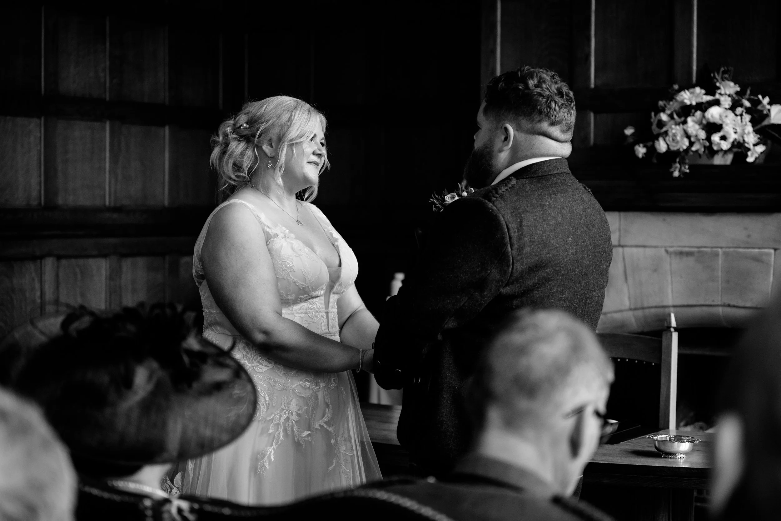 A bride and groom exchanging vows during a wedding ceremony, holding hands, inside a wood-paneled room with floral decorations.