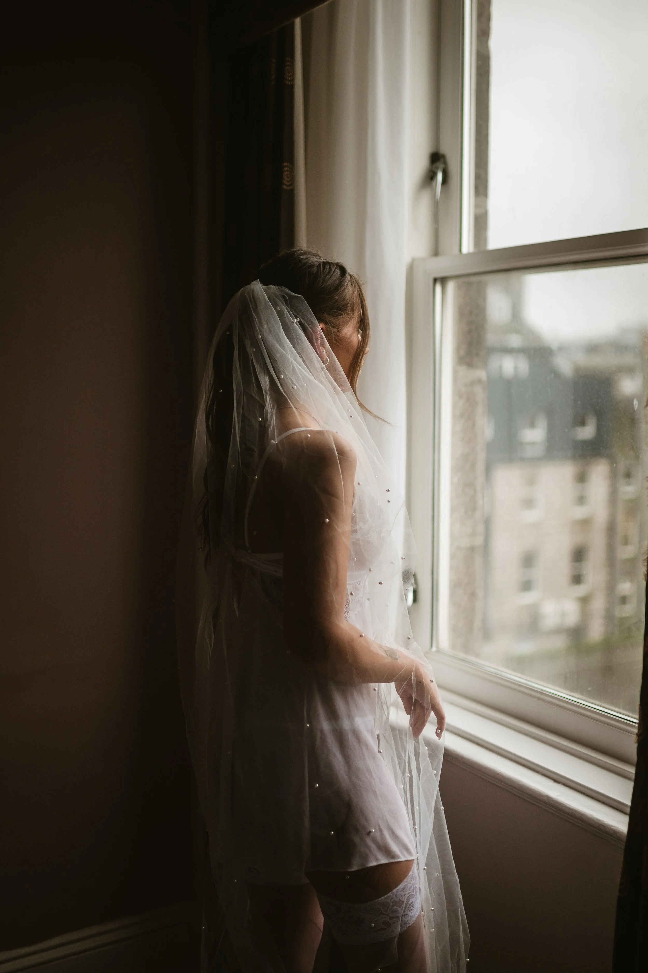 A woman in bridal lingerie, stockings, and a veil, looking out of a window in a dimly lit room.