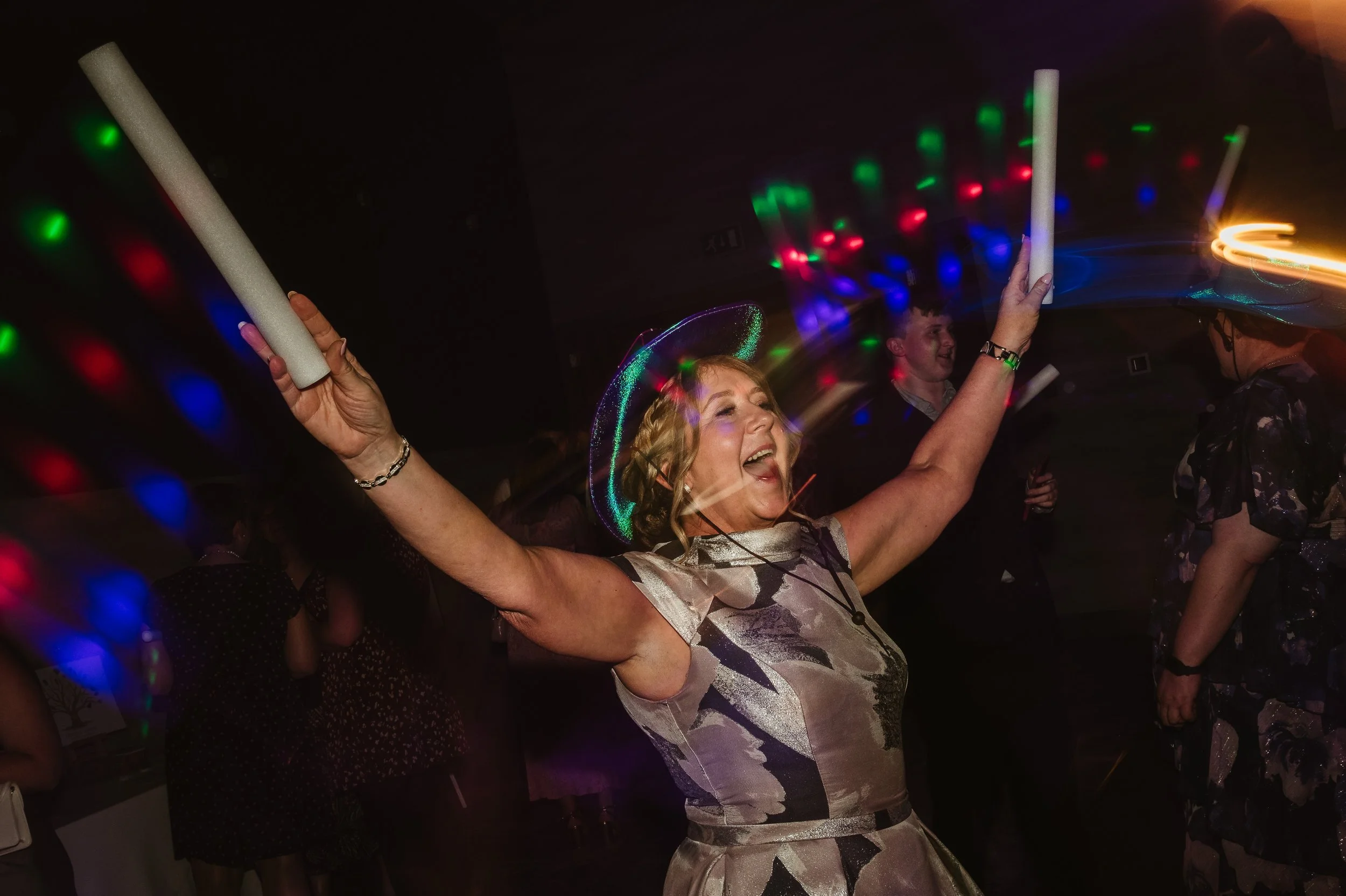 Woman celebrating and dancing at a party, holding glow sticks with colorful lights around her, wearing a floral dress and a glittery hat, with other people in the background.