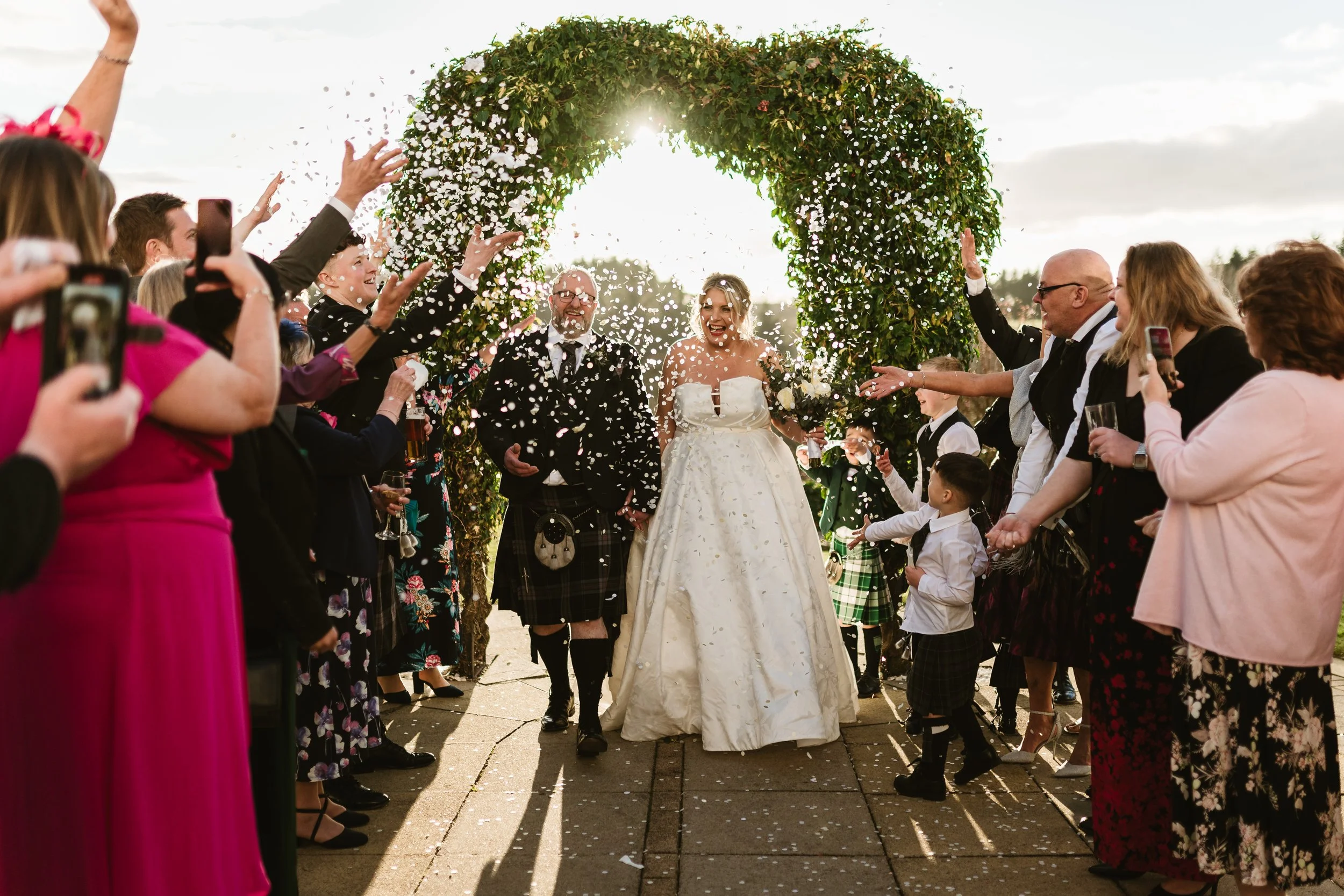 A bride and groom walking through a celebratory archway made of greenery and flowers, surrounded by guests cheering and taking photos, with confetti falling around them during a wedding ceremony outdoors in the late afternoon.