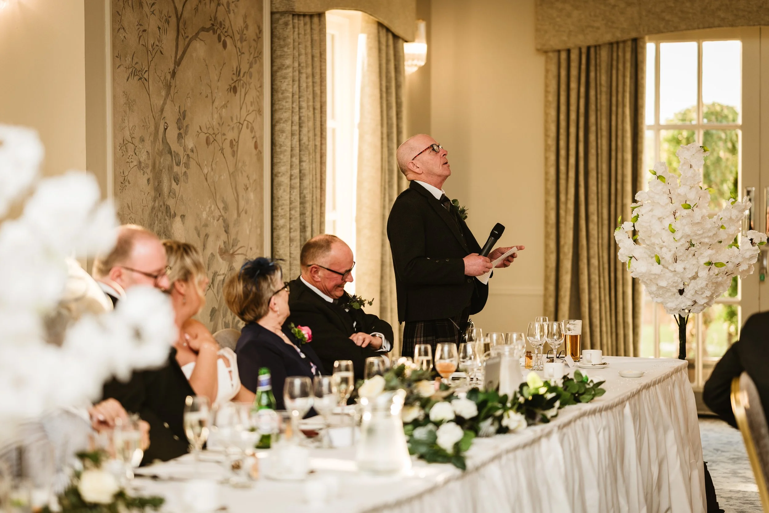 A man in a black tuxedo giving a speech at a wedding reception, holding a microphone and a paper, standing at a decorated head table with guests seated along it. The room has elegant curtains and a large window with daylight outside.
