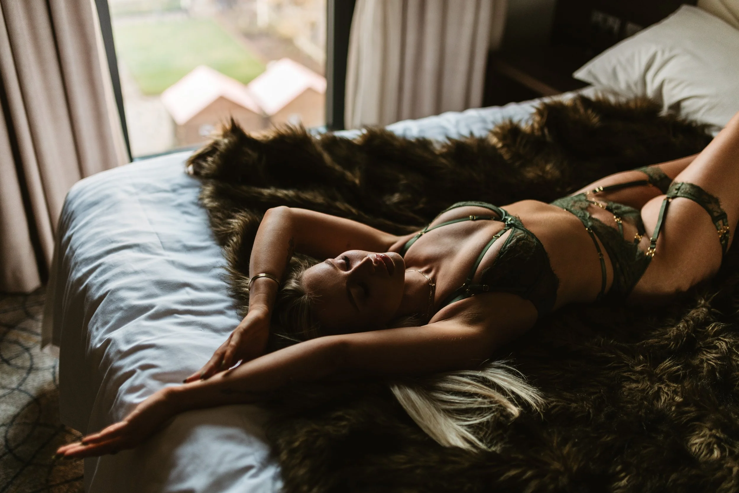 A woman in green lingerie lying on a bed with her eyes closed, stretching with her arms above her head, beside a large black dog.