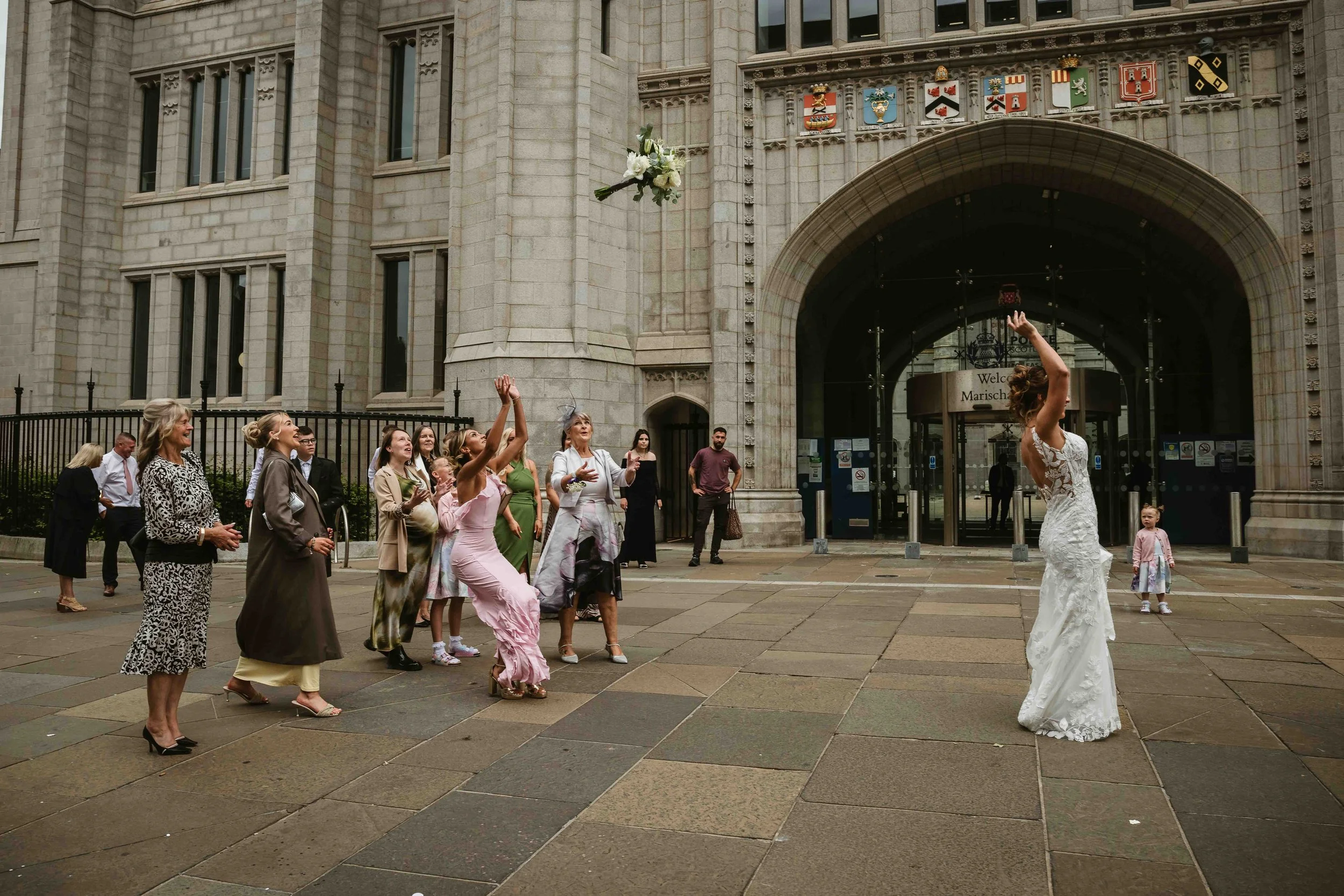 A bride throws her bouquet to a group of women and girls outside a large stone building, likely a church or government building, during a wedding celebration.