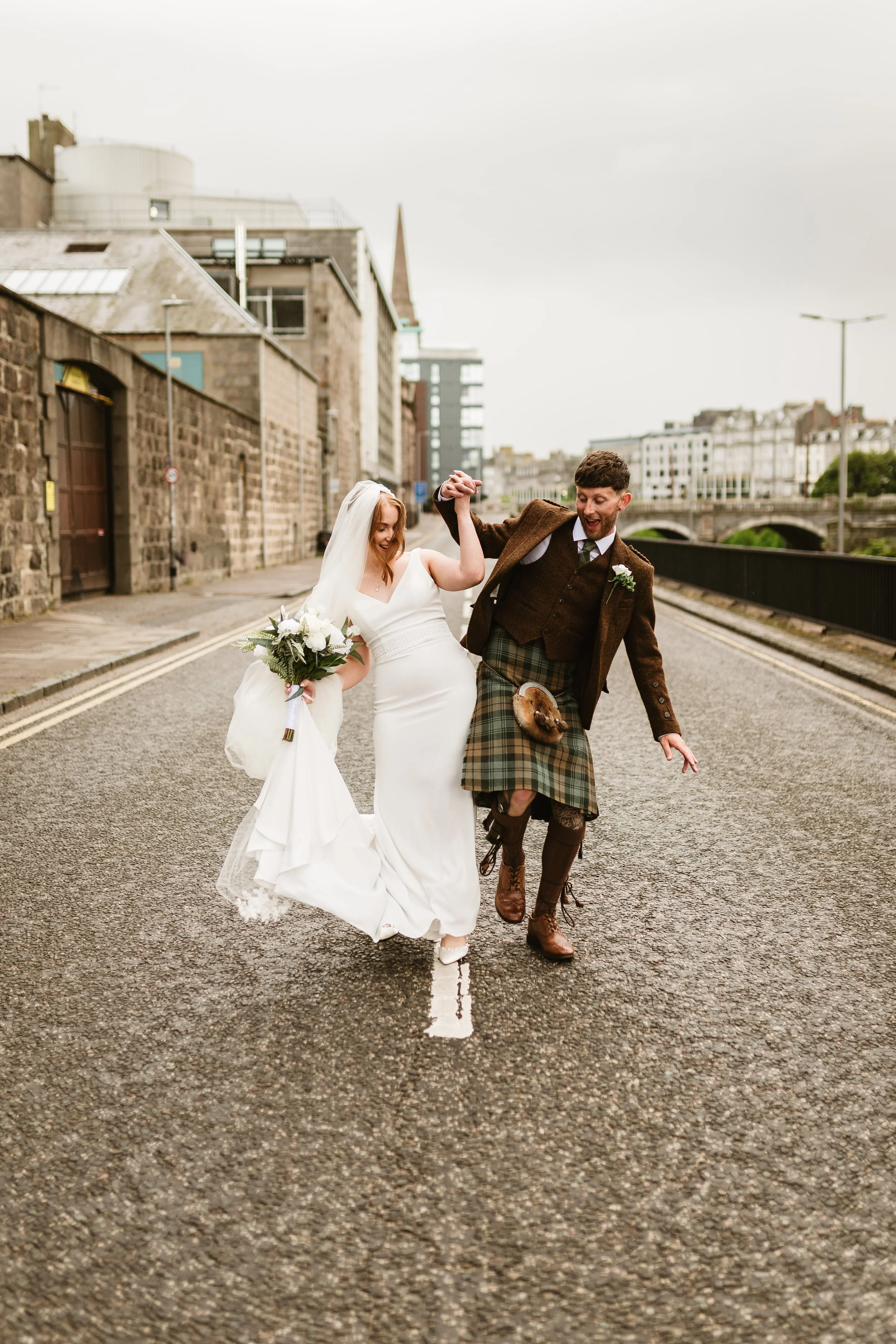 A bride in a white wedding dress and a groom in a traditional Scottish kilt and jacket are dancing together on a city street.