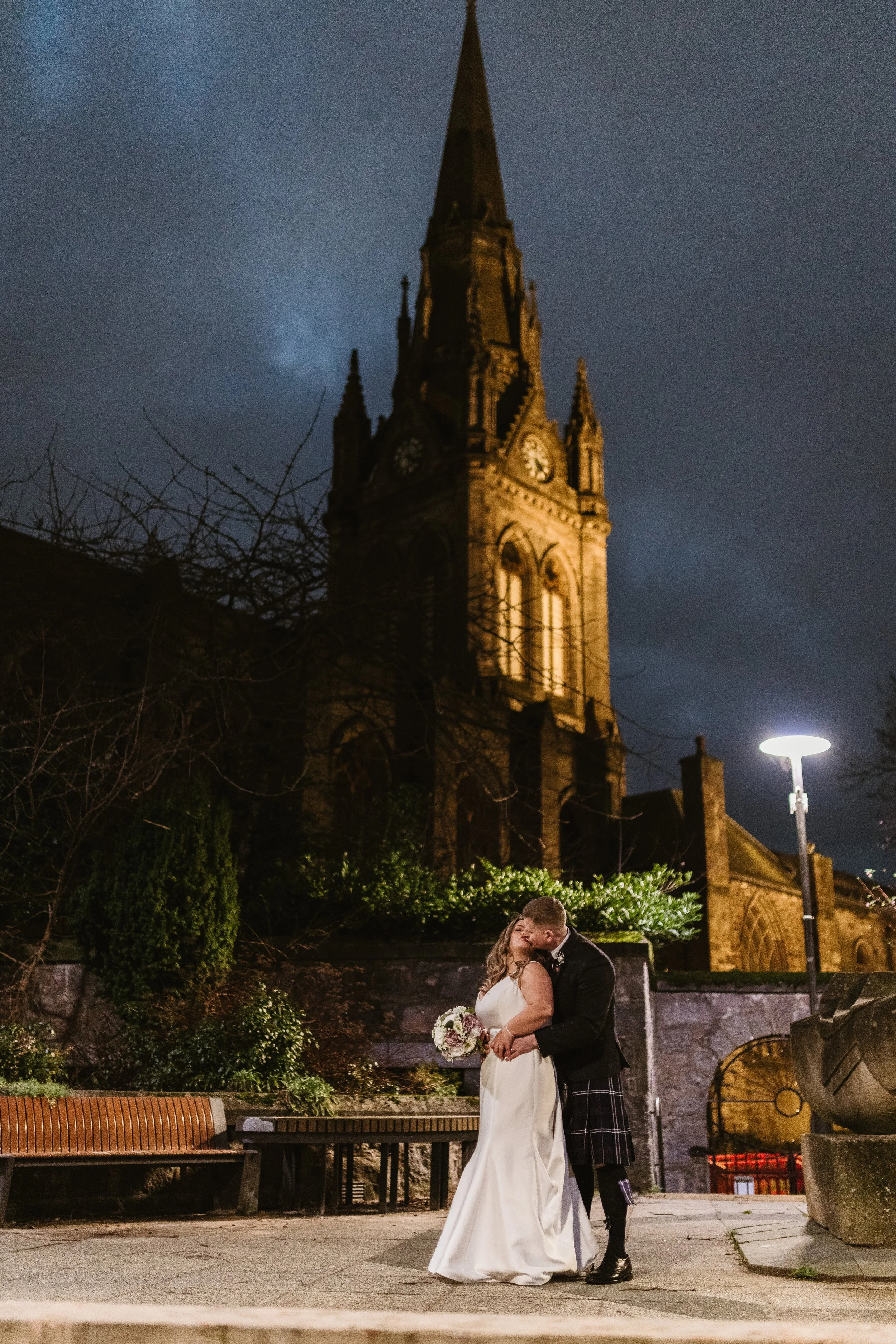 Bride and groom sharing a kiss outdoors at night under a large illuminated church with a tall spire.