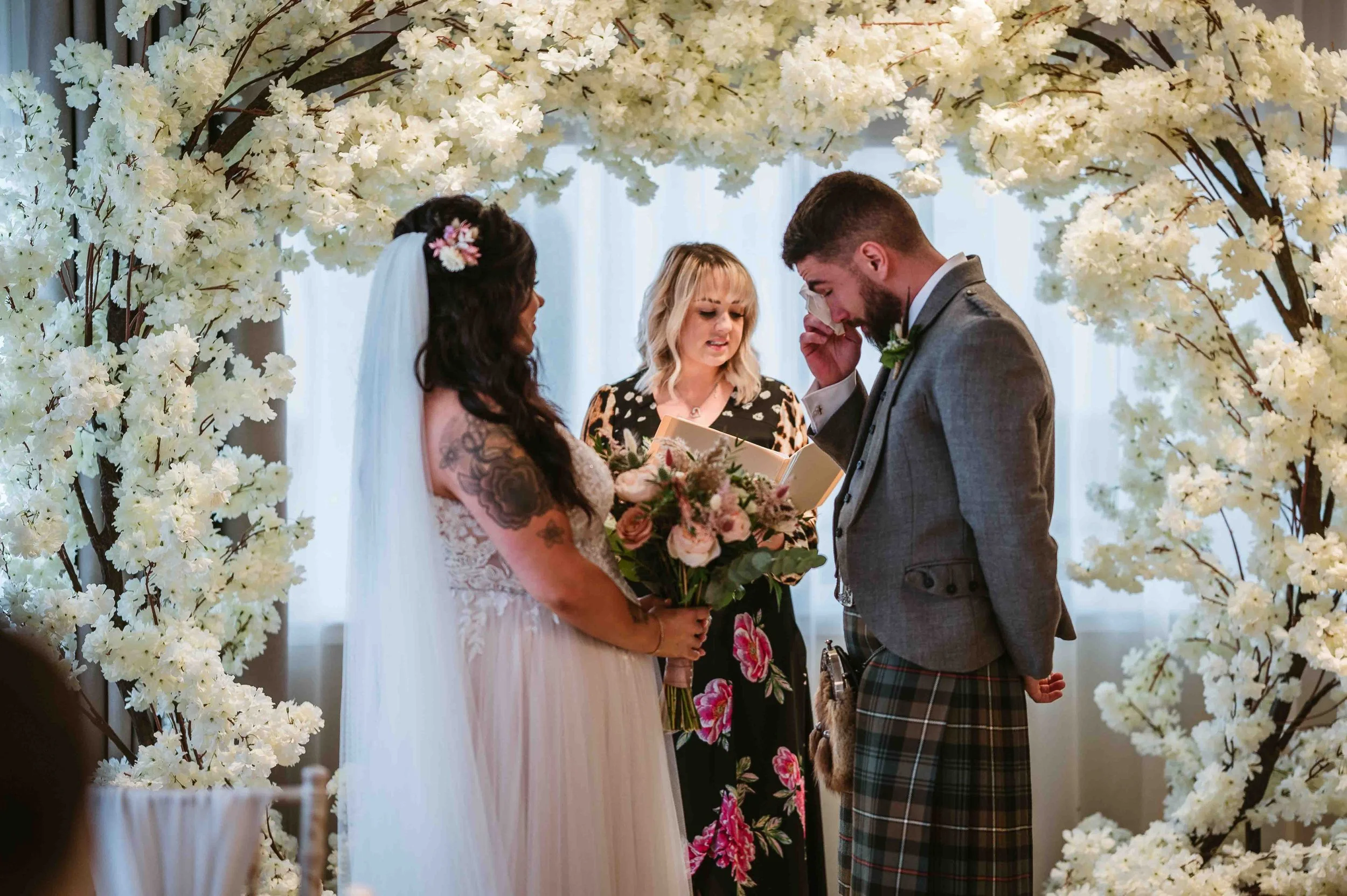 A wedding ceremony with a bride and groom standing under a floral arch, the bride holding a bouquet and the officiant reading from a book.