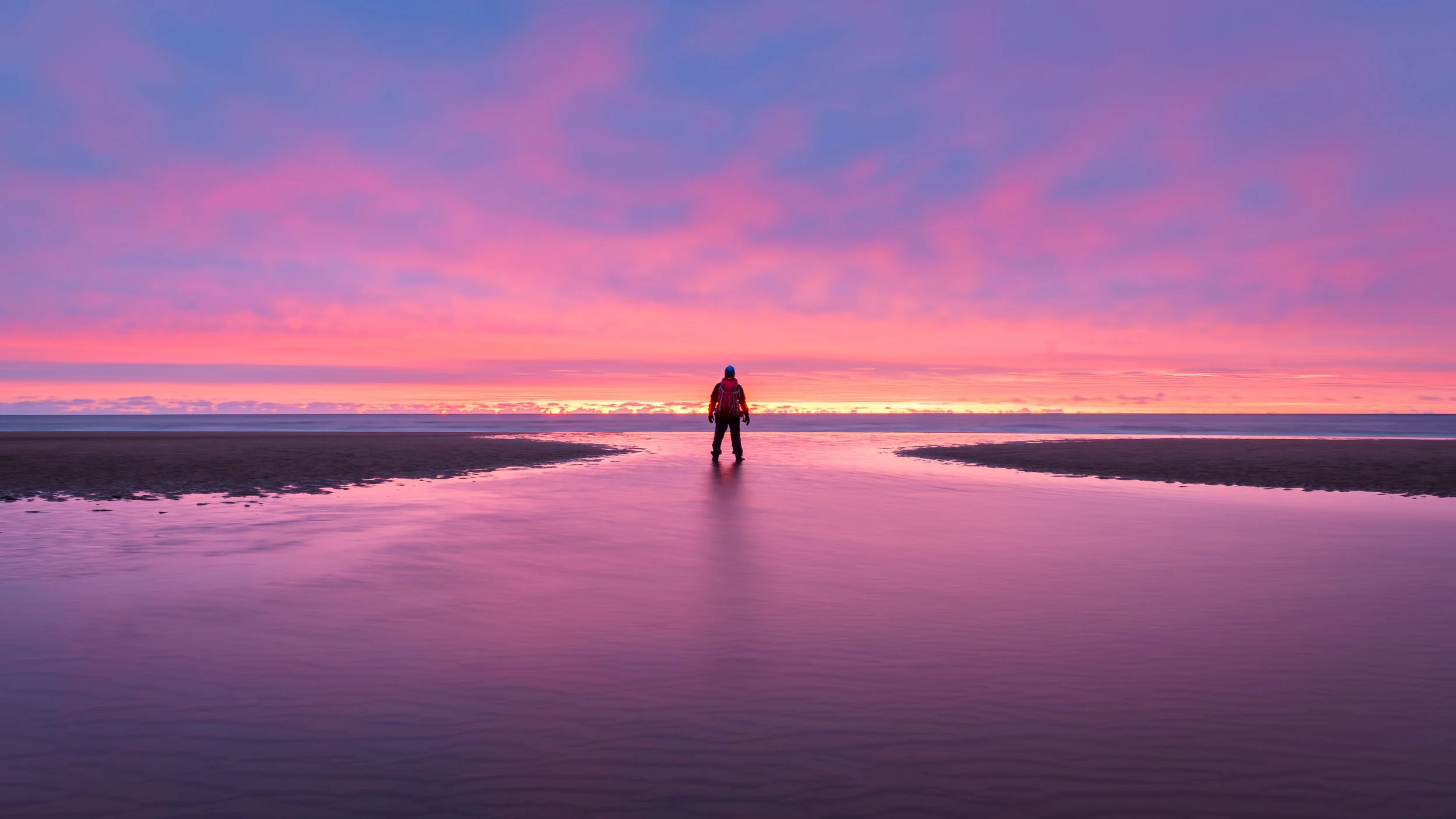Person standing on a beach at sunset or sunrise, with pink and purple clouds reflecting on the water, and the distant ocean horizon in the background.