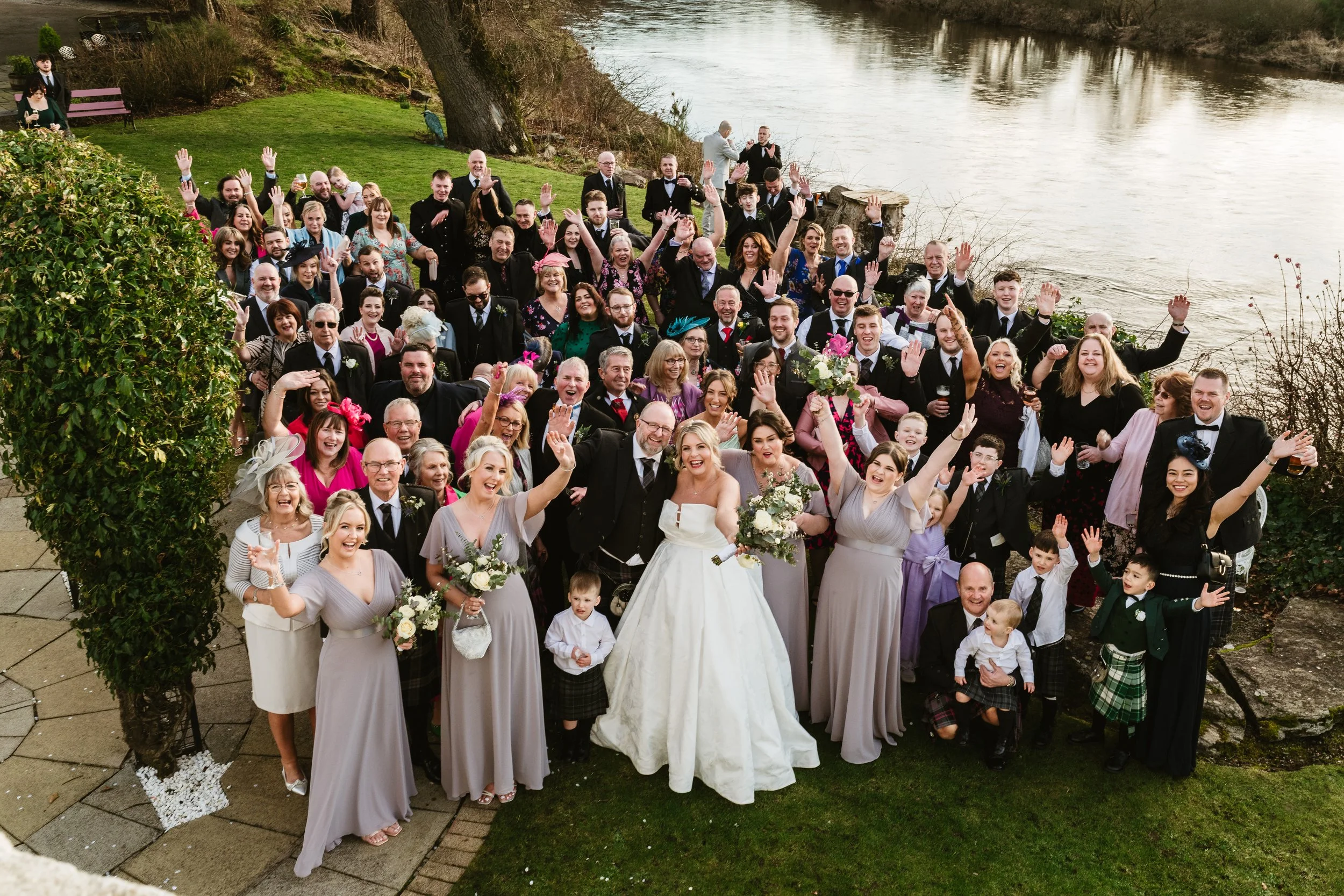 A large wedding group outdoors by a river, with the bride and groom in the center, surrounded by family and friends, all smiling and waving.