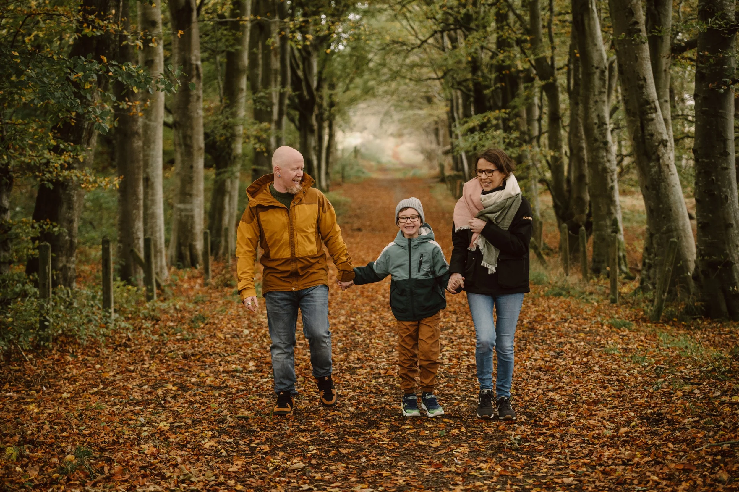 A family of three, a father, mother, and young boy, walking hand in hand on a leaf-covered trail in an autumn forest, smiling and enjoying the walk.