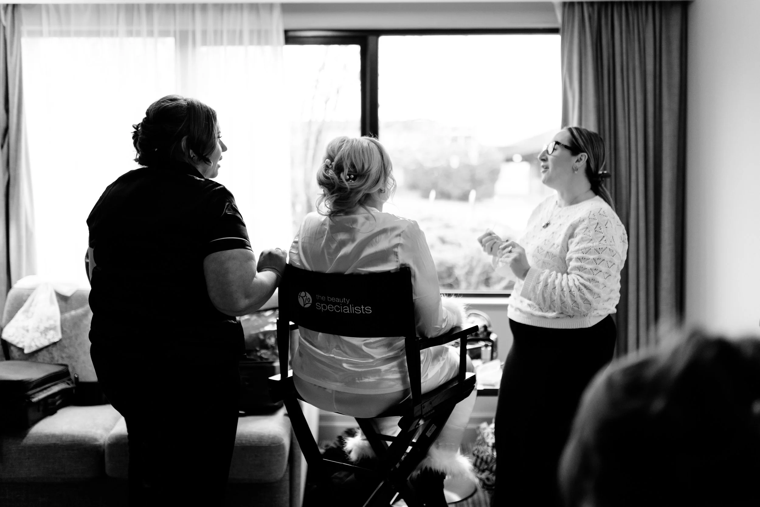 Three women in a room, one seated in a chair, while the other two stand, engaged in conversation near a large window with curtains.