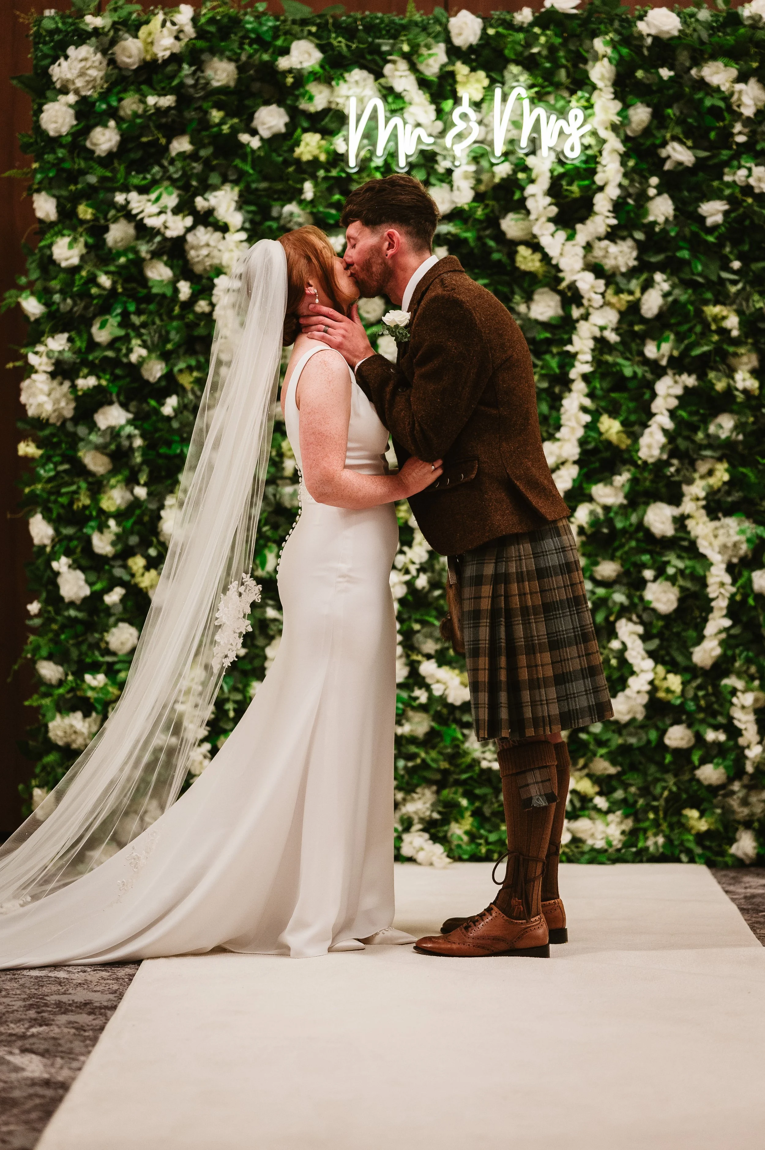 A bride and groom sharing a kiss during their wedding ceremony with a backdrop of white flowers and greenery, a neon sign reading 'Mrs. & Mr.' behind them.