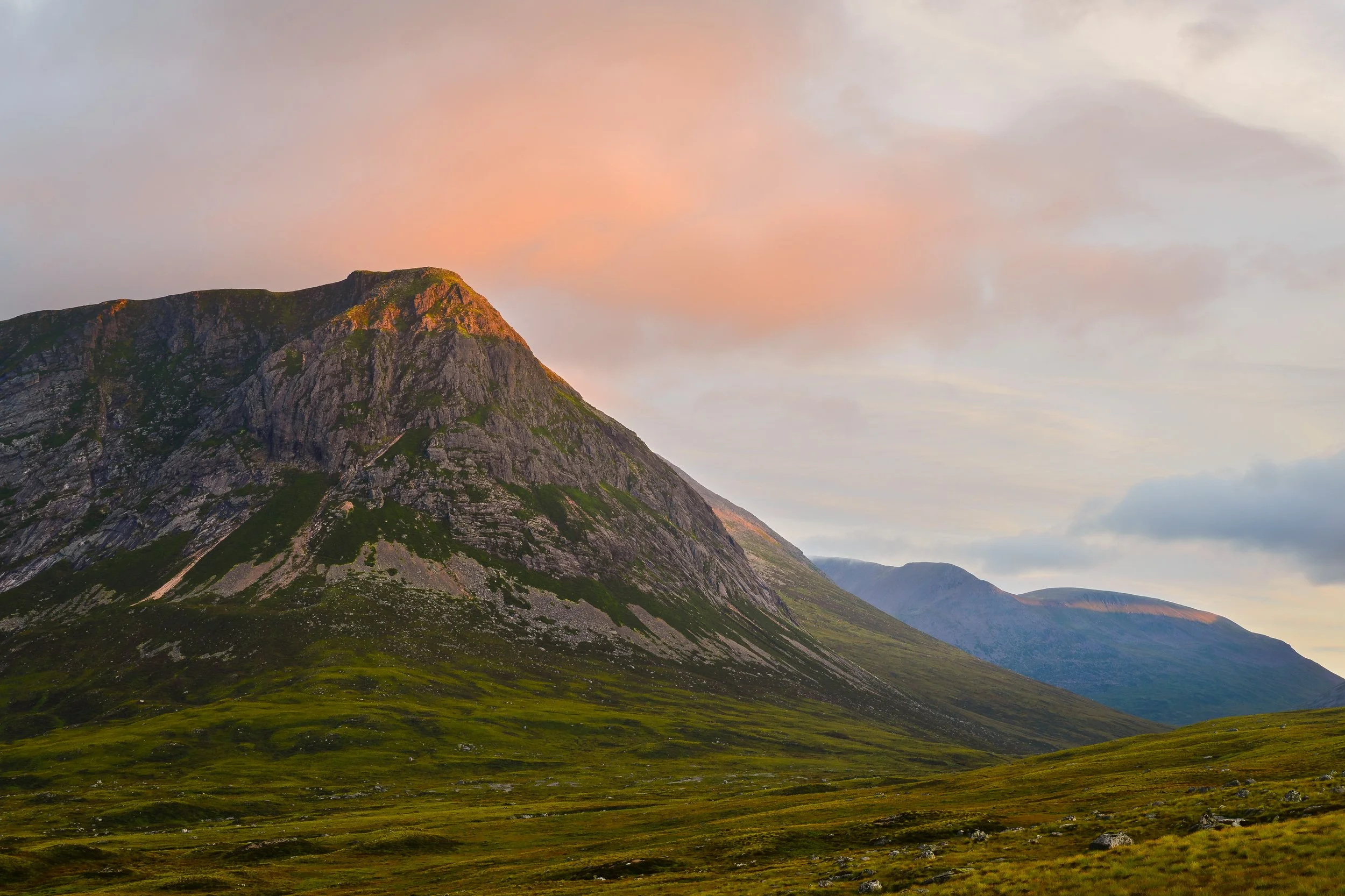 A mountain landscape at sunset with green grassy foreground, rocky slopes, and pink-tinged clouds in the sky.