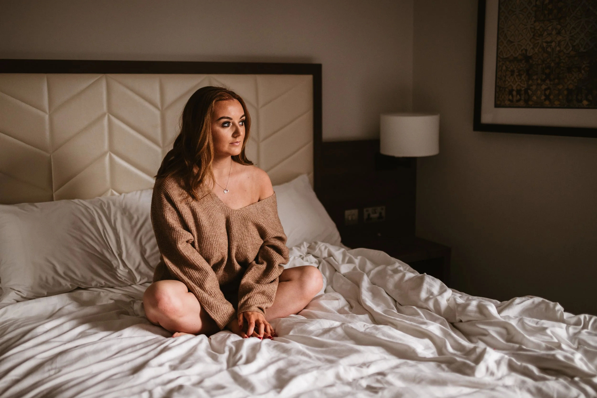 A woman with long light brown hair sits cross-legged on a bed in a hotel room, looking thoughtfully to the side. She is wearing a loose brown sweater and a jewelry necklace. The bed has white sheets and a padded headboard, with a nightstand and lamp 