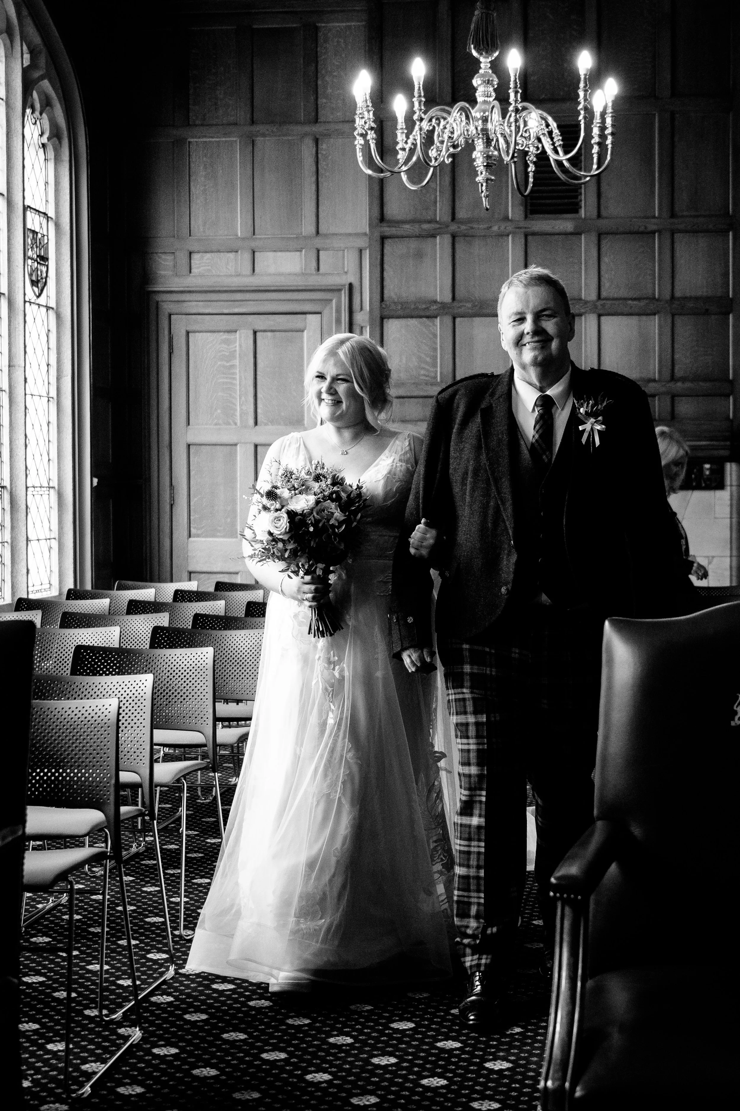 A black and white photograph of a bride walking down the aisle with a man in a formal setting, possibly a church or ceremony hall, with wooden paneling and a chandelier overhead.