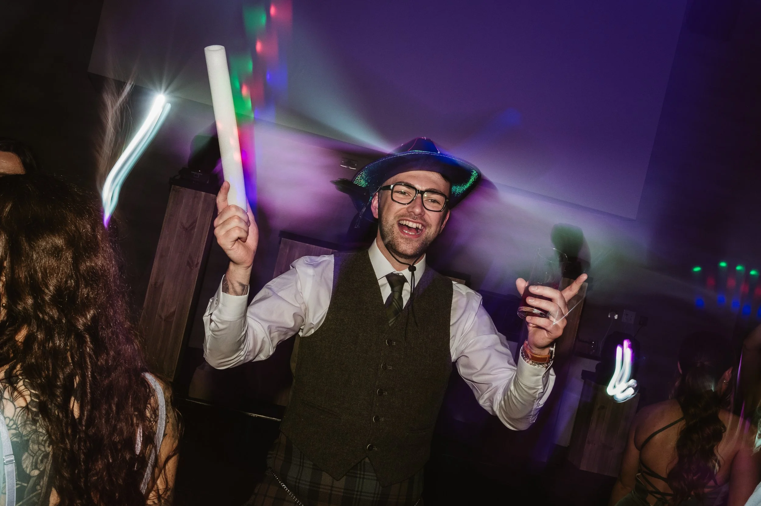 A man wearing glasses, a vest, and a hat dancing and holding a drink at a party with colorful lights.