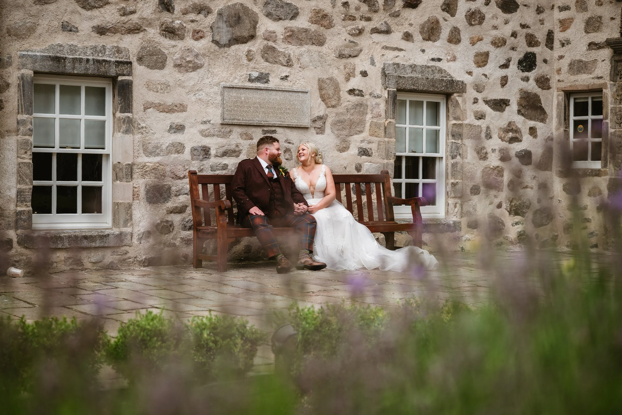 A bride and groom sit on a wooden bench against a stone wall, holding hands and smiling at each other. The bride is wearing a white wedding dress with lace details, and the groom is dressed in a dark suit with a tartan kilt. There are two windows beh
