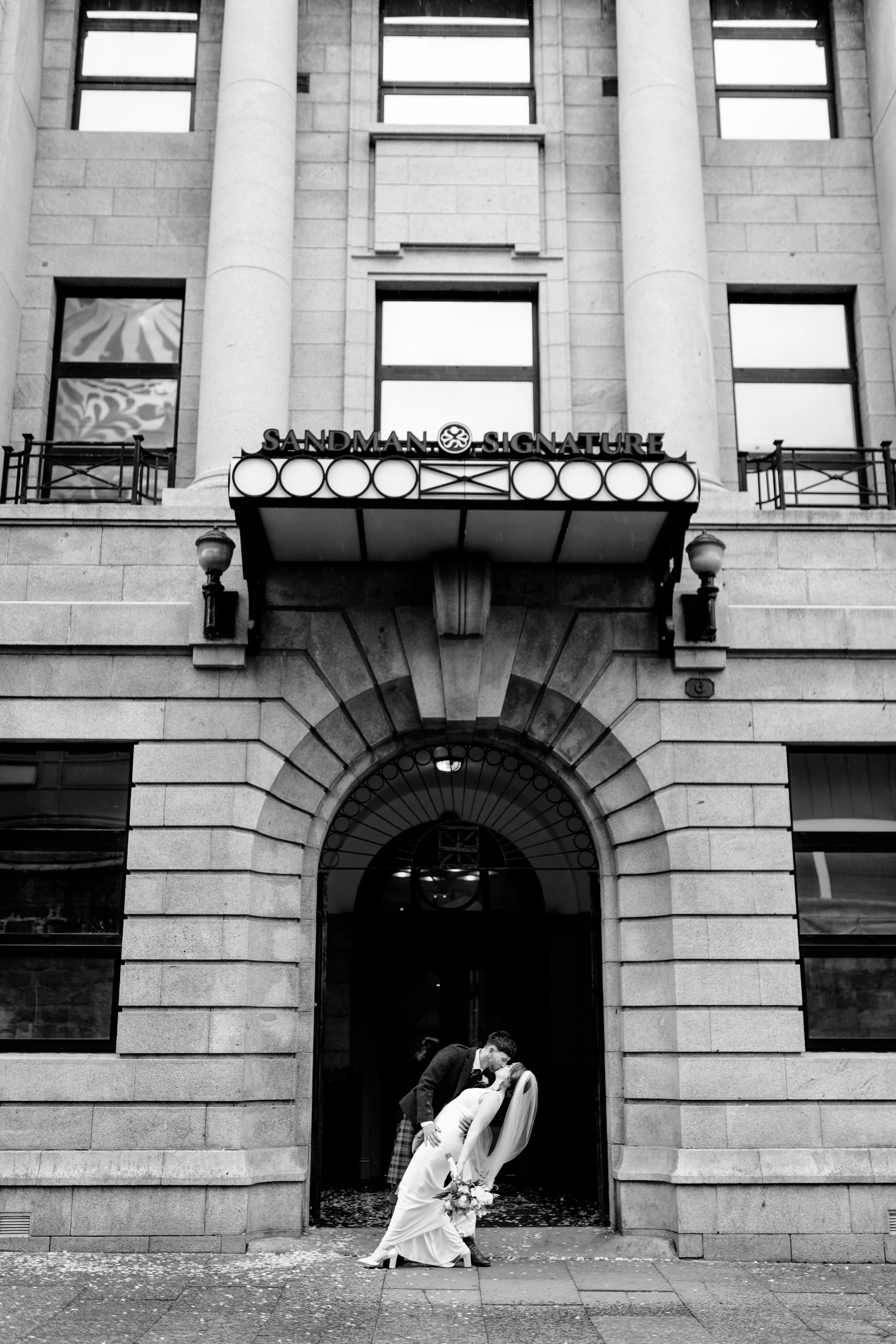 A wedding couple sharing a kiss in front of a building with the sign 'Sandman Signature' above the entrance.