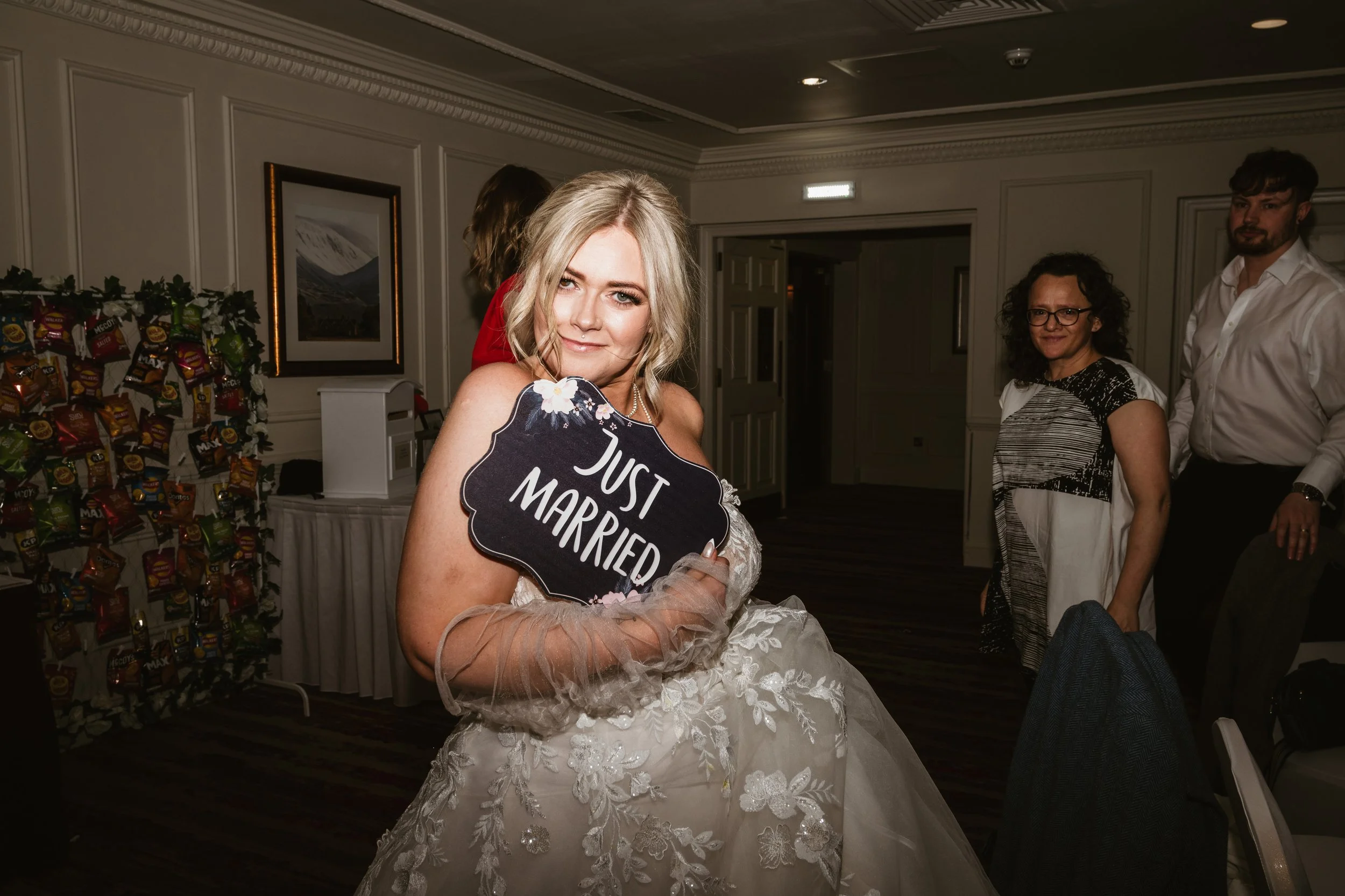 A bride in a wedding dress holding a sign that says 'Just Married' at a wedding reception.