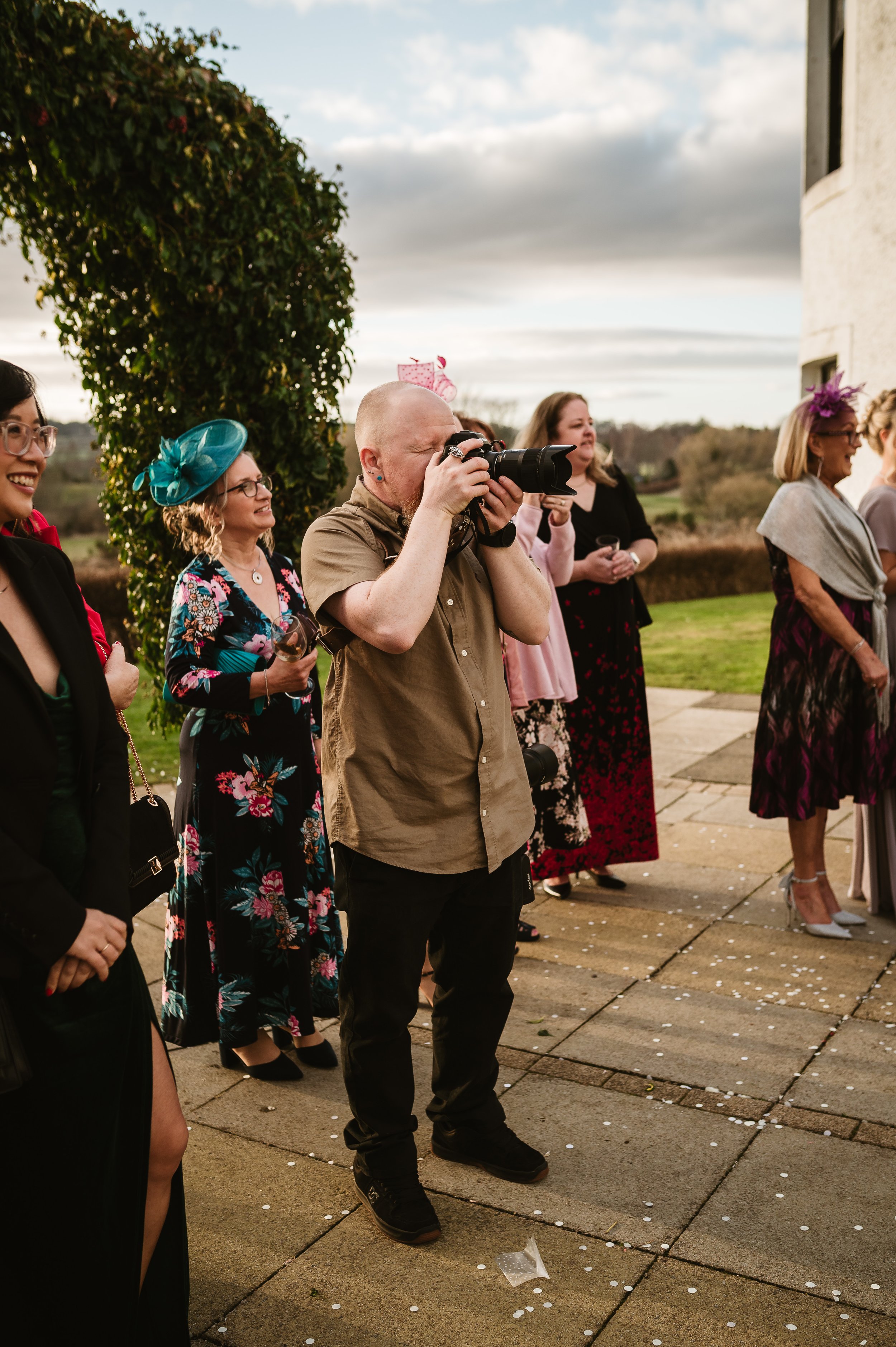 A photographer taking a picture at an outdoor event with several people dressed in colorful attire, on a patio with a scenic landscape in the background.