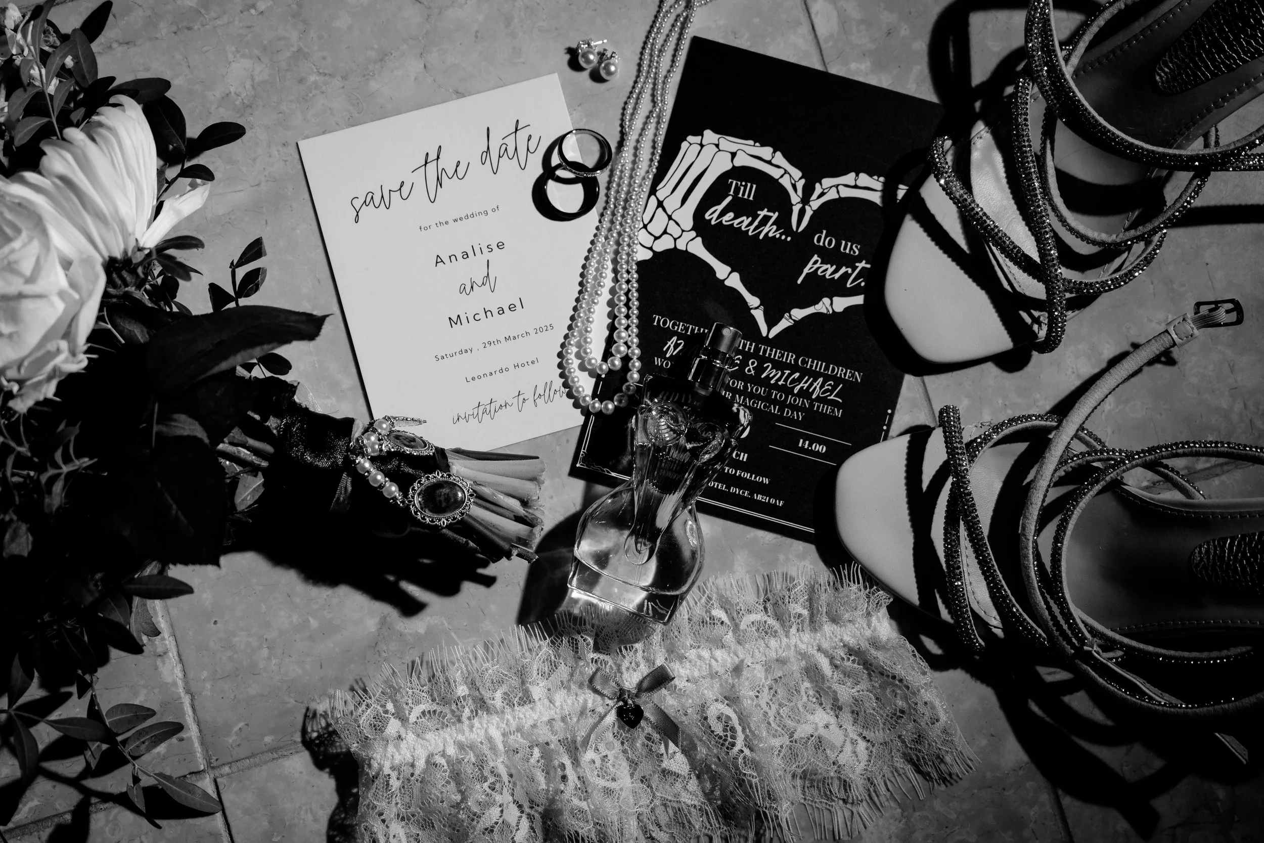 Wedding invitation, jewelry, perfume bottle, high-heeled shoes, flowers, lace garter, and other accessories arranged on a flat surface in a black-and-white photo.