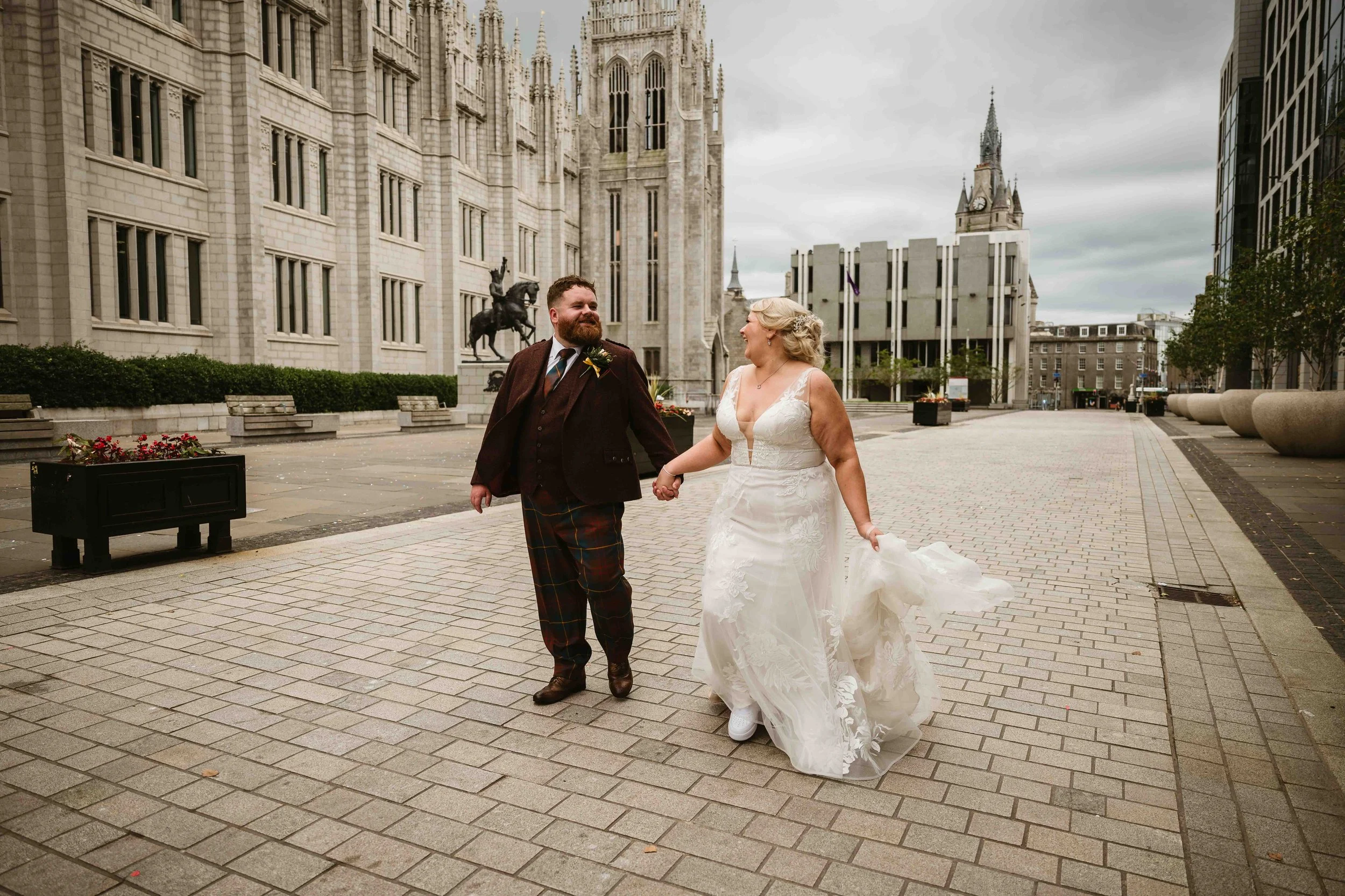 A newlywed couple holding hands and smiling while walking on a city street with historic and modern buildings, including a church and a clock tower, in the background.