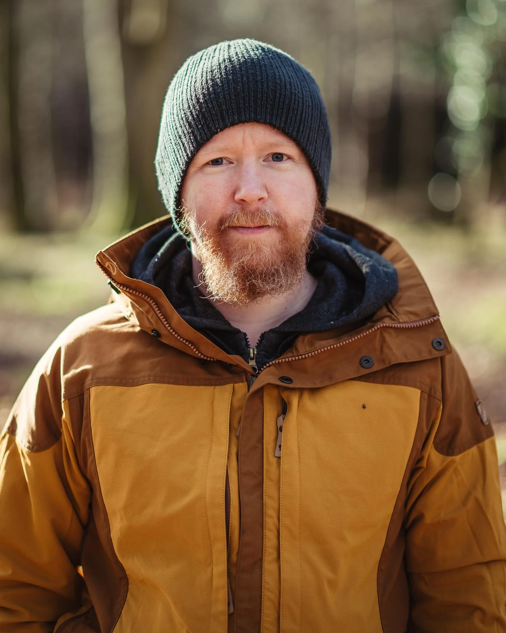 Man with a red beard wearing a dark blue knit beanie and brown jacket outdoors in a forest.