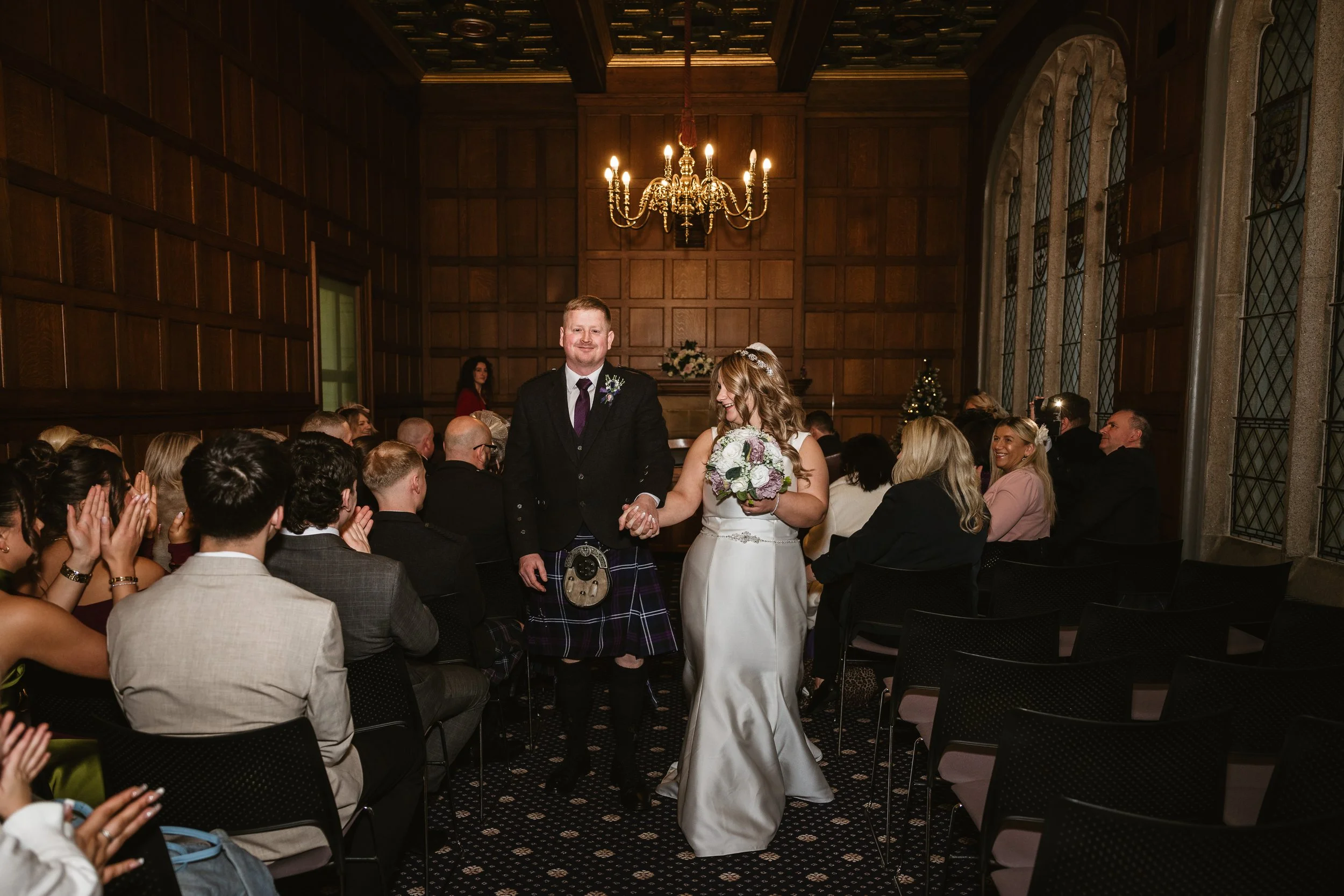 A bride and groom walking hand in hand down the aisle after getting married in a wood-paneled room with a chandelier overhead. The bride is holding a bouquet of flowers, and the groom is wearing a kilt and a suit. Guests are seated and clapping on ei