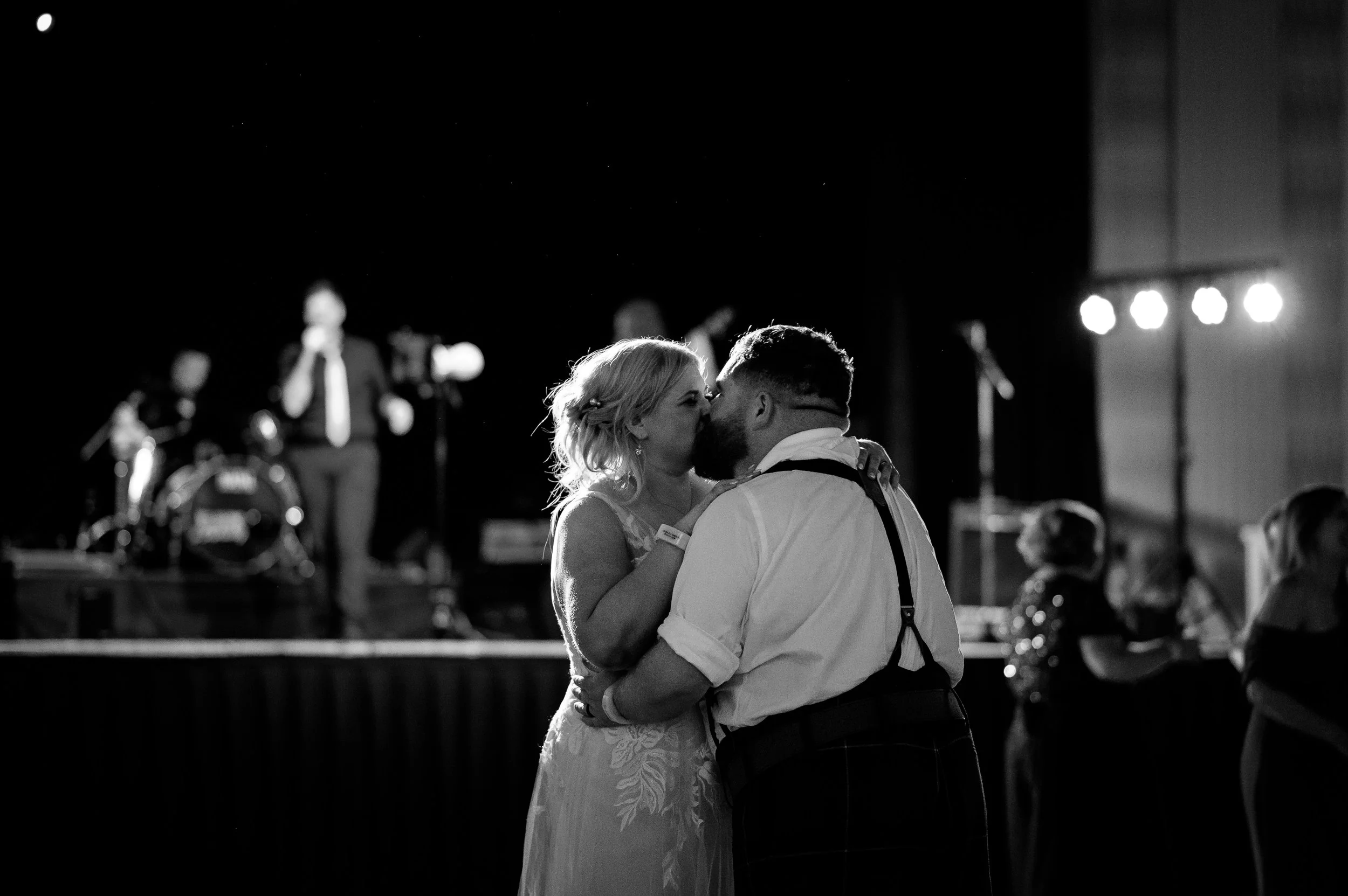 A couple dancing closely at a wedding reception, with a band playing live music on stage in the background.