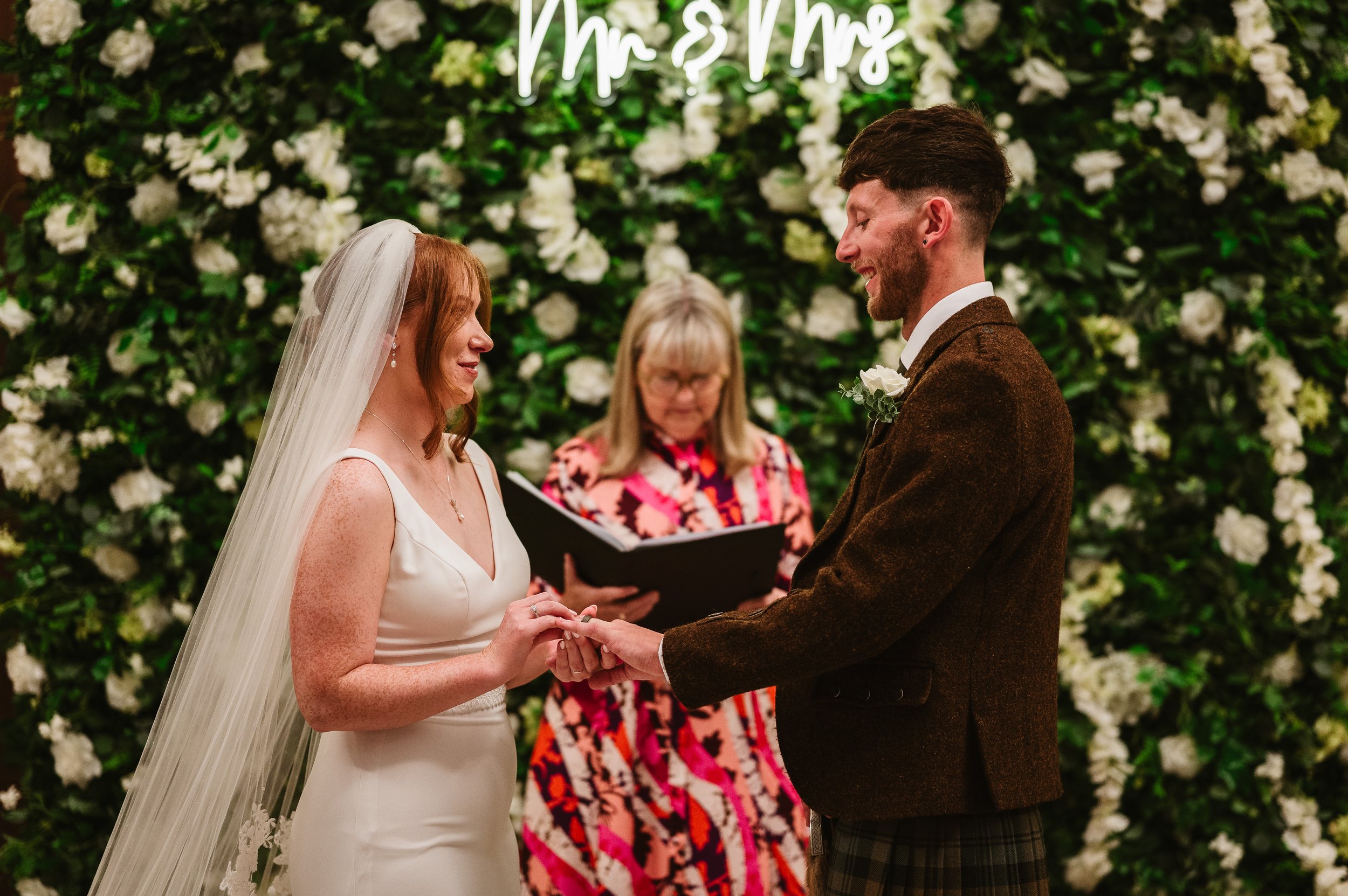 A bride and groom exchanging rings during their wedding ceremony, with a floral backdrop and officiant overseeing the process.