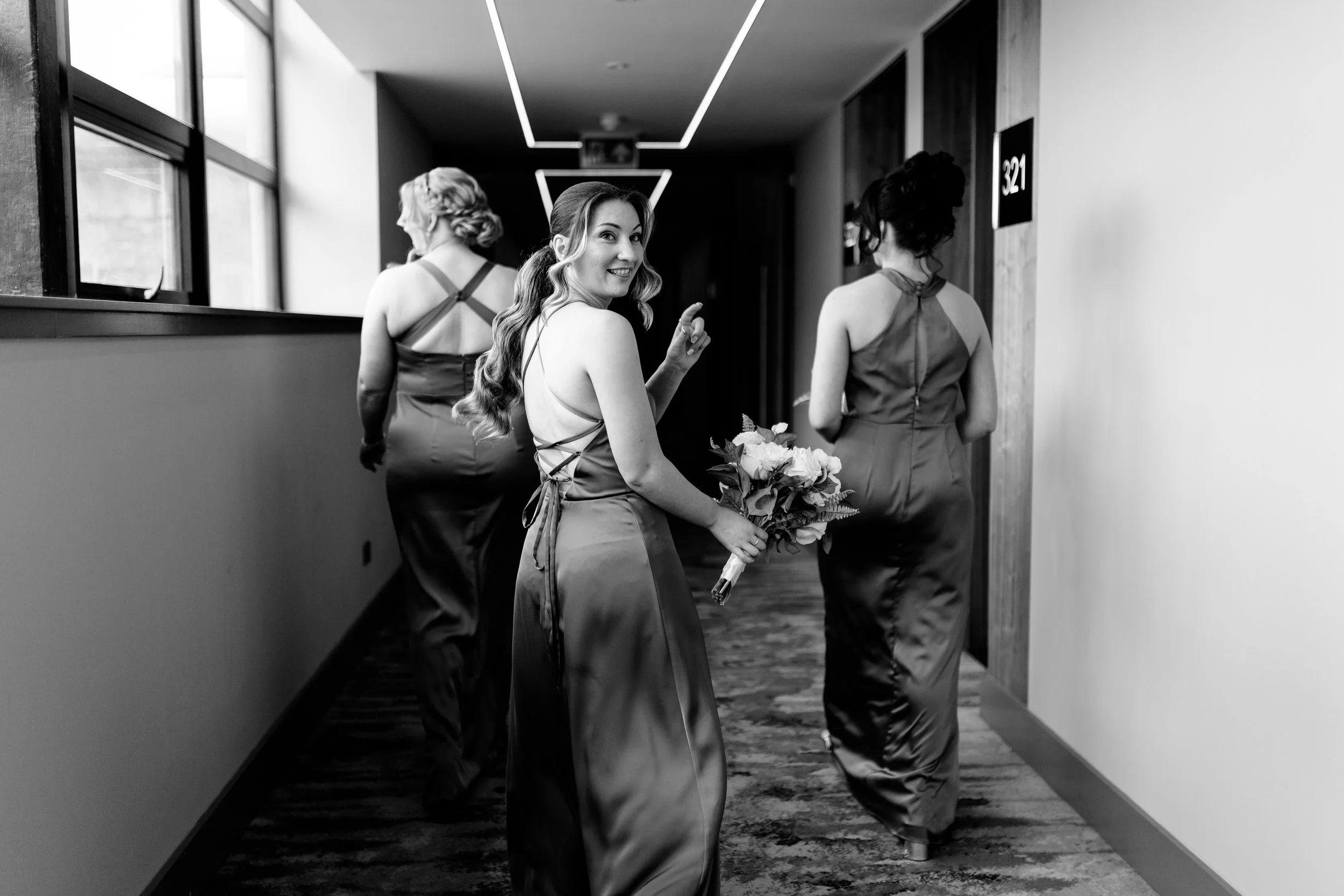 Three women in satin dresses walking down a hallway, one holding a bouquet and smiling at the camera.