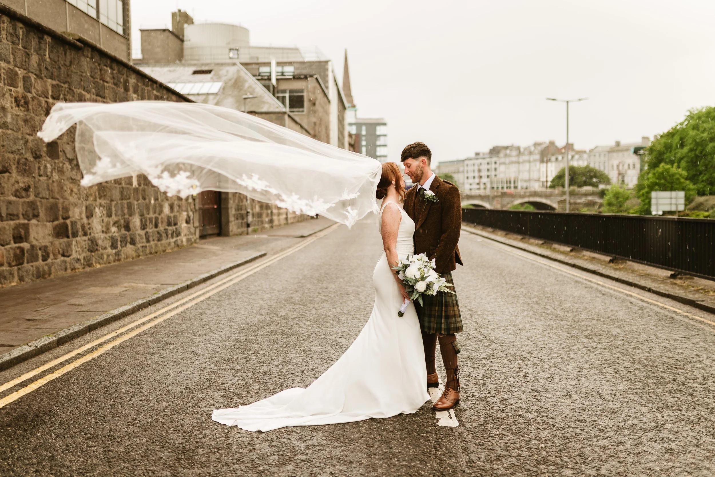 A bride and groom standing close together on a city street, looking at each other, with the bride holding a bouquet and the groom wearing a kilt. The bride's veil is flying in the wind.