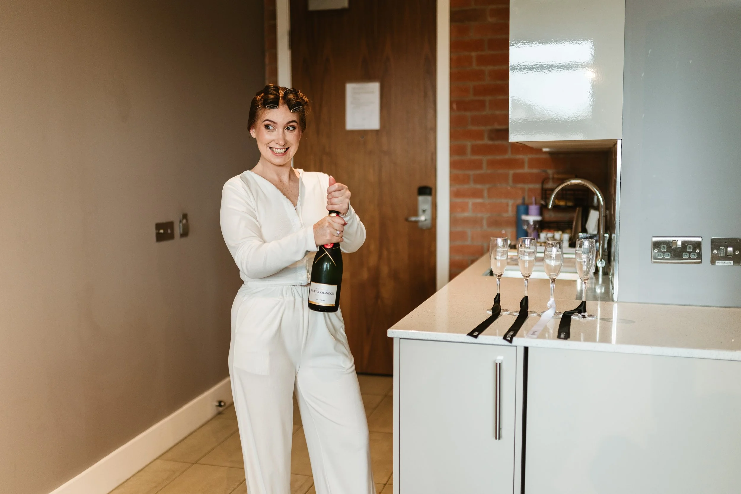 A woman in white pajamas opening a bottle of champagne in a modern kitchen with glasses of champagne on the counter.