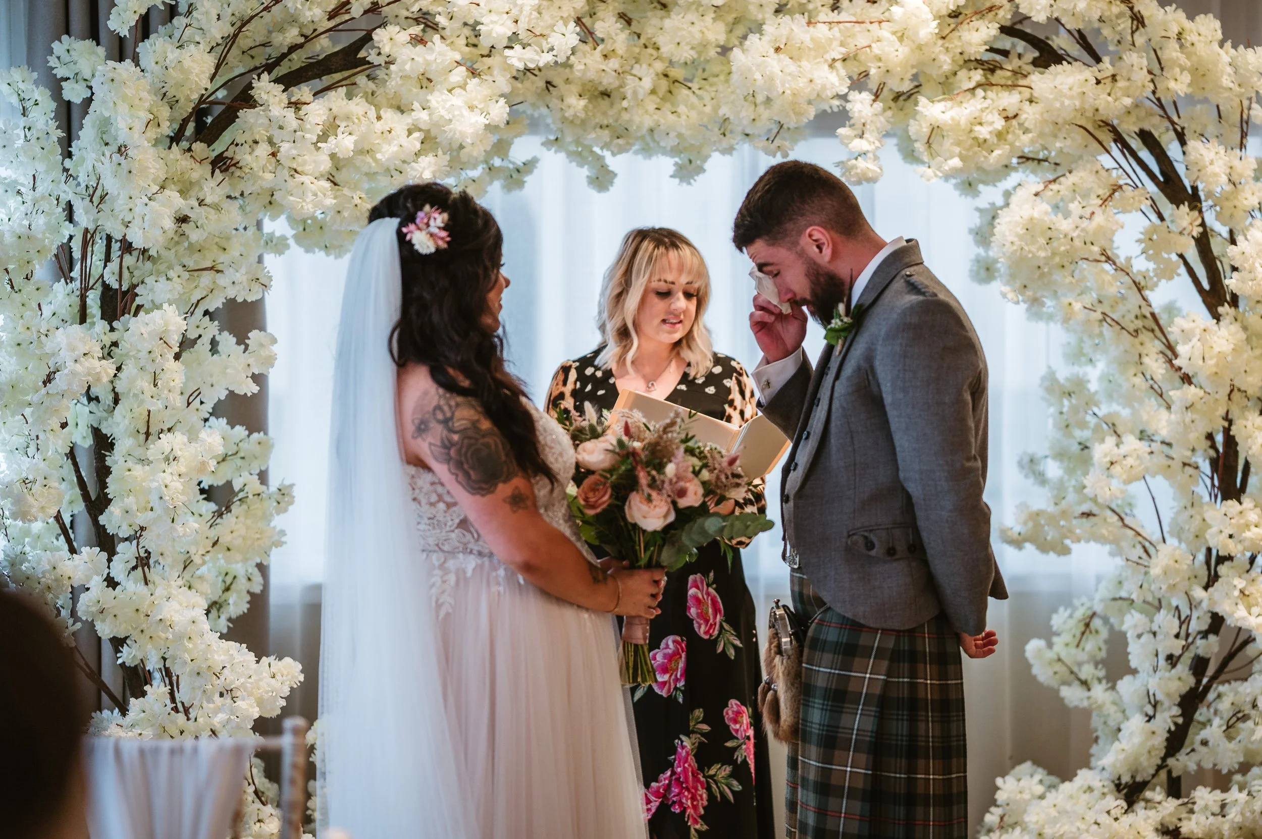 A wedding ceremony with a bride and groom standing under a floral arch, exchanging vows, with an officiant holding a book.
