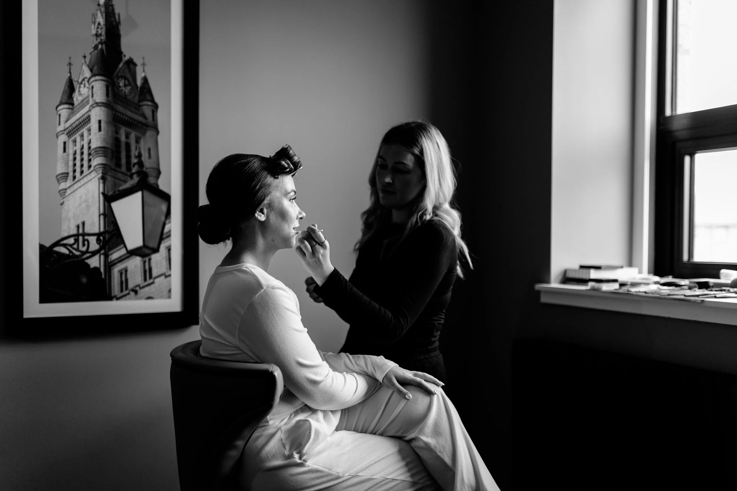 A woman in a white dress sits on a chair while a makeup artist applies makeup to her face in a well-lit room with a window and framed artwork on the wall.