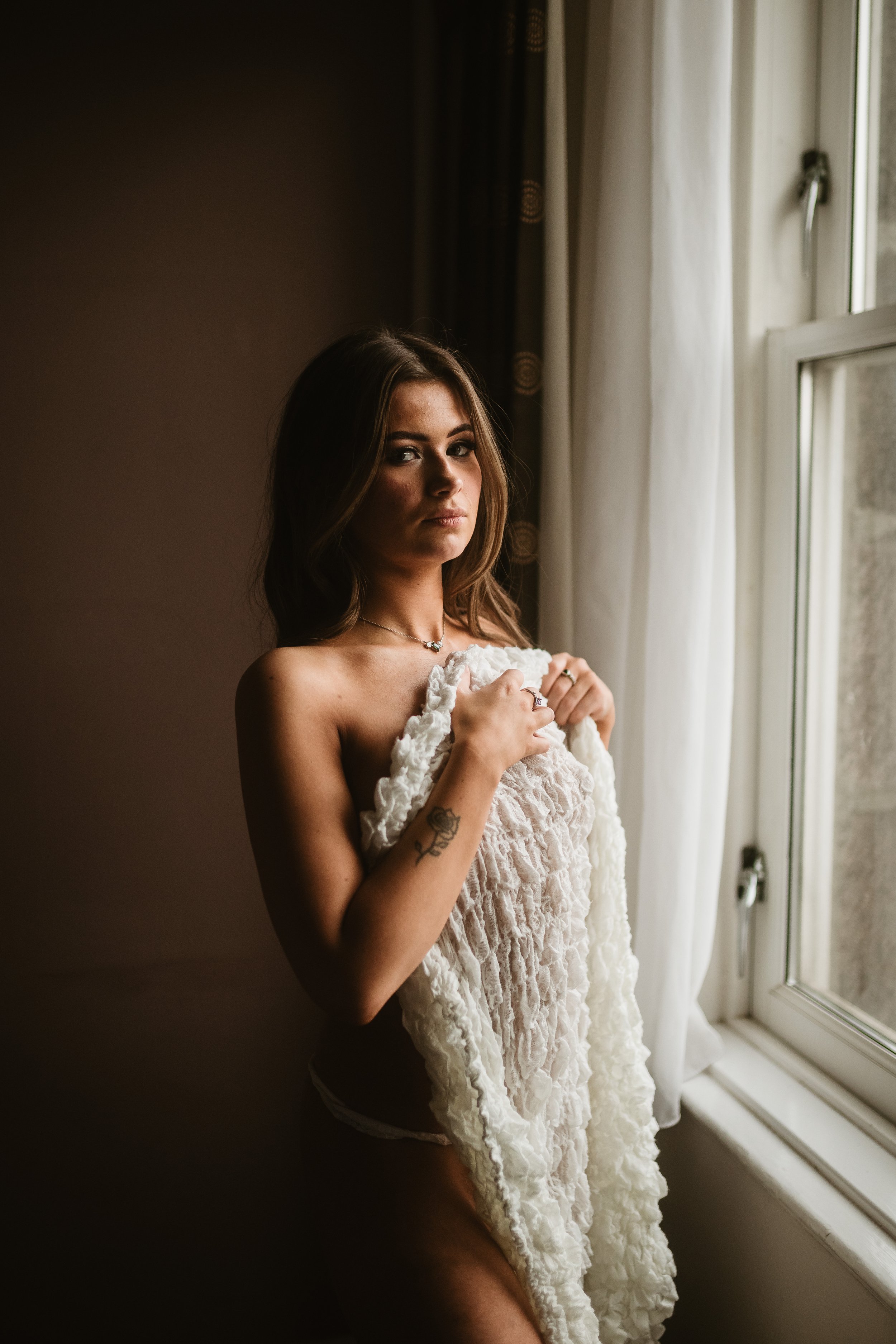 A young woman standing by a window, holding a soft, textured white blanket against her chest, with natural light illuminating her face and upper body.